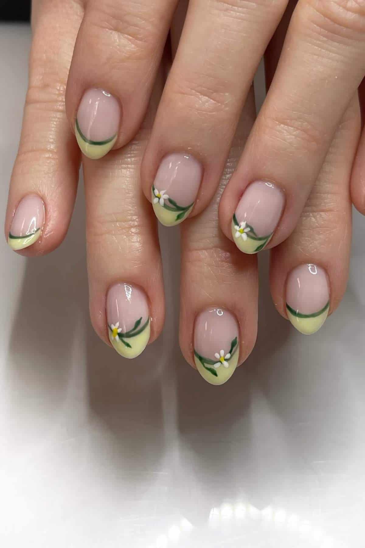 A close-up of hands with a manicure featuring pale yellow French tips and small white floral designs with green leaves on each fingernail.