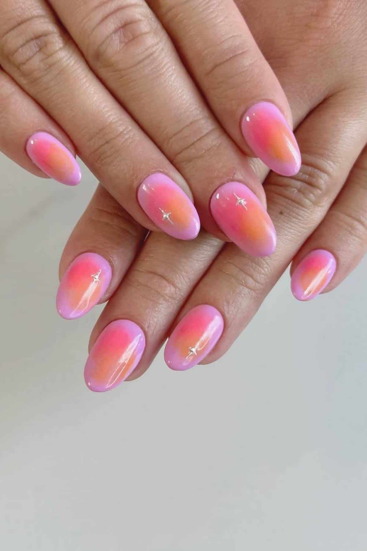 Hands with almond-shaped nails featuring a pink to orange gradient ombre design and small star-shaped accents on a light background.