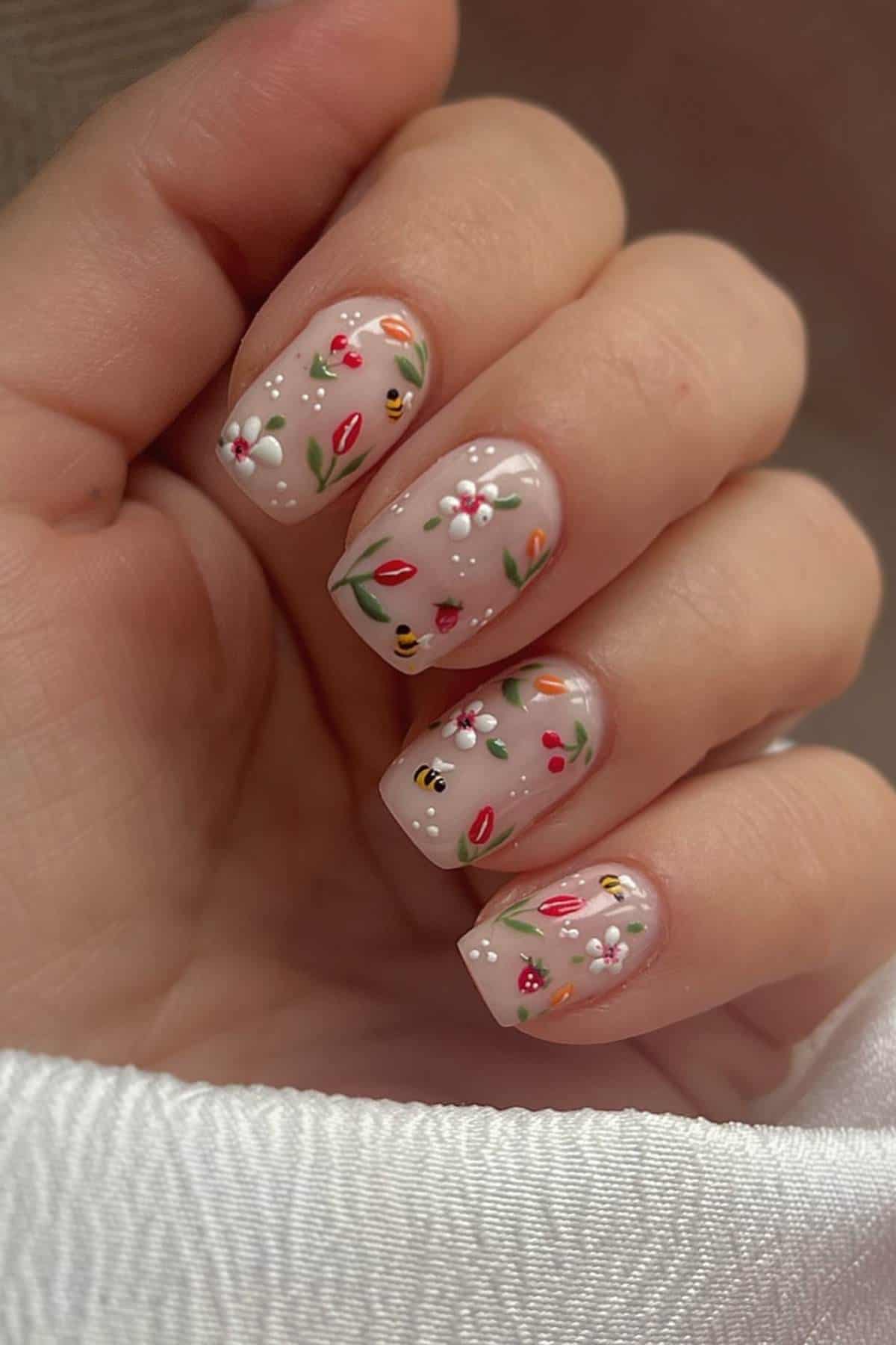 A hand with short, square-shaped nails featuring intricate floral nail art with small bees, red and white flowers, and green leaves on a nude base.