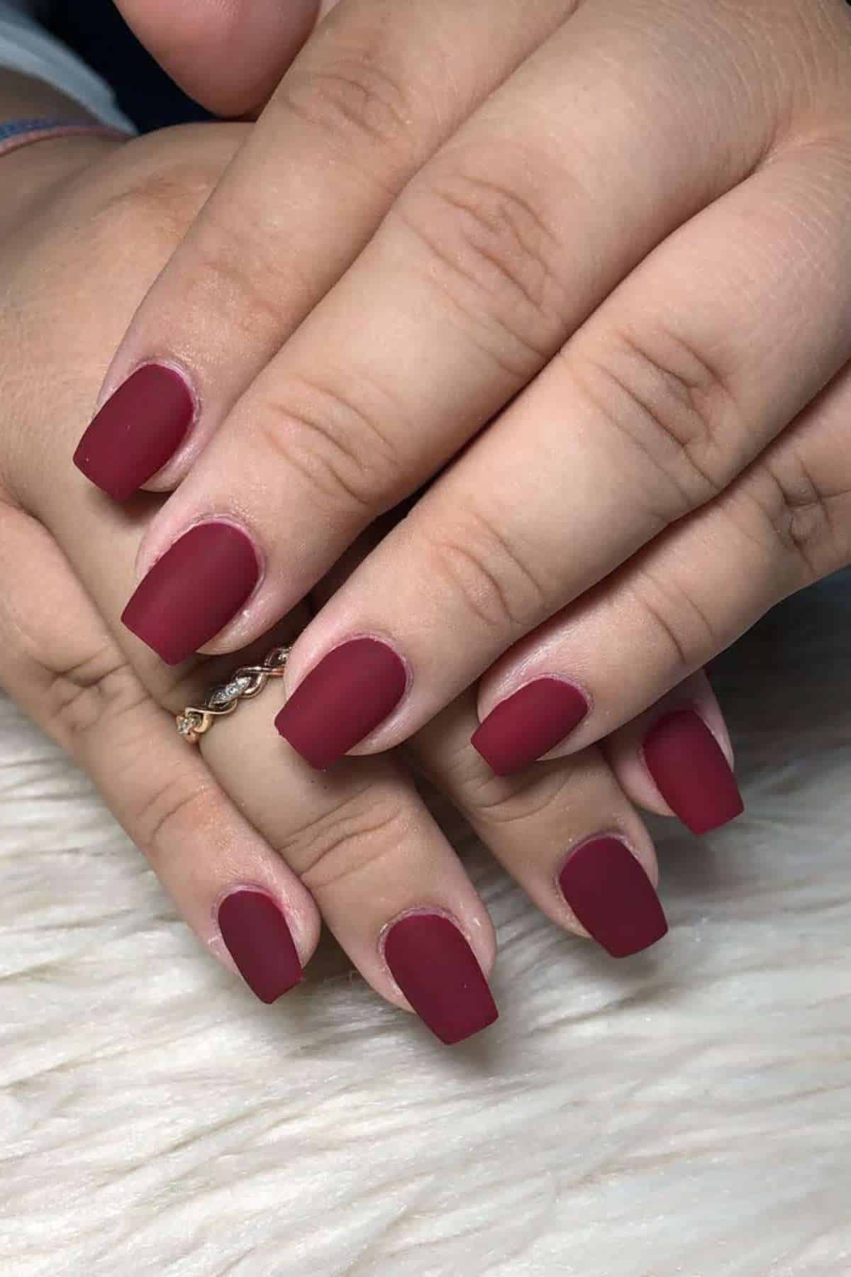 Close-up of hands with short, square-shaped fingernails painted in a matte burgundy color, resting on a light textured surface.
