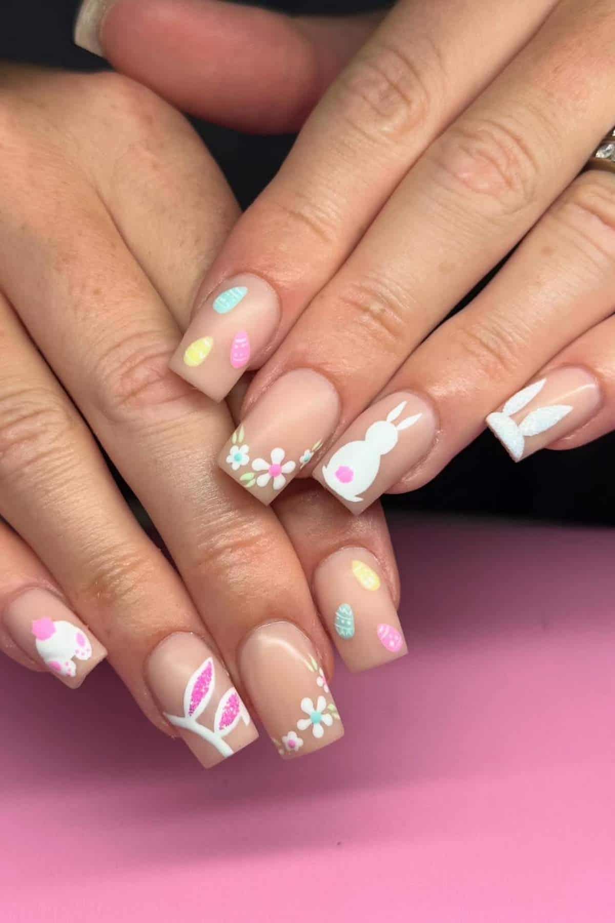 A close-up of hands with nude-colored nails decorated with Easter-themed designs, including white bunnies, colorful eggs, flowers, and leaves.