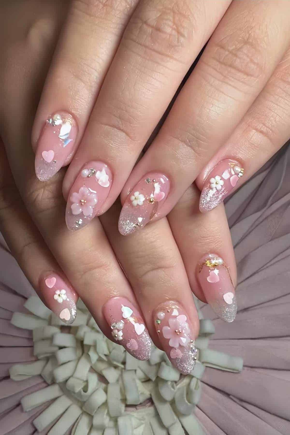 Hands with almond-shaped nails, painted pink with glitter tips, decorated with 3D white flowers, pink hearts, and small rhinestones, displayed over a pale fabric background.