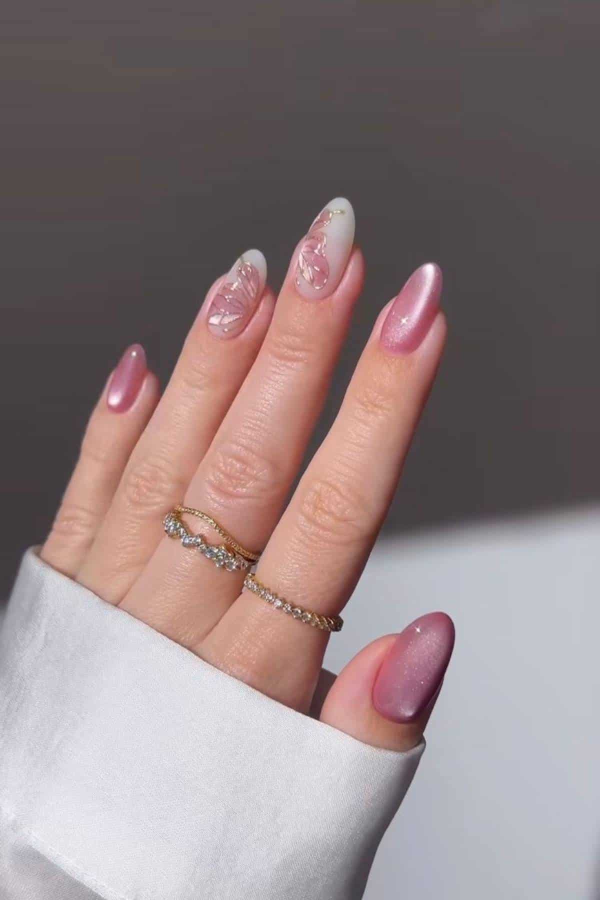 A hand with long, almond-shaped pink nails, featuring one accent nail with white and pink floral nail art. The person is wearing two gold rings and a white sleeve.