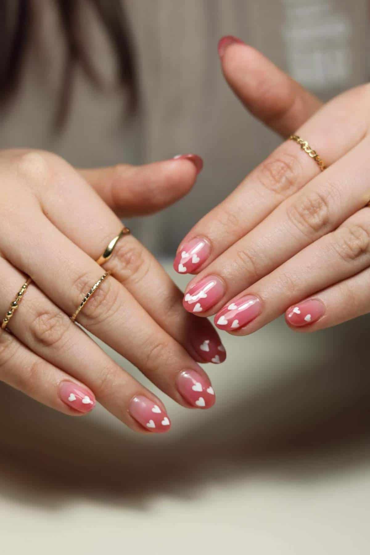 Hands with almond-shaped nails feature a pink base and white heart designs on the tips. The person is wearing simple gold rings on several fingers.