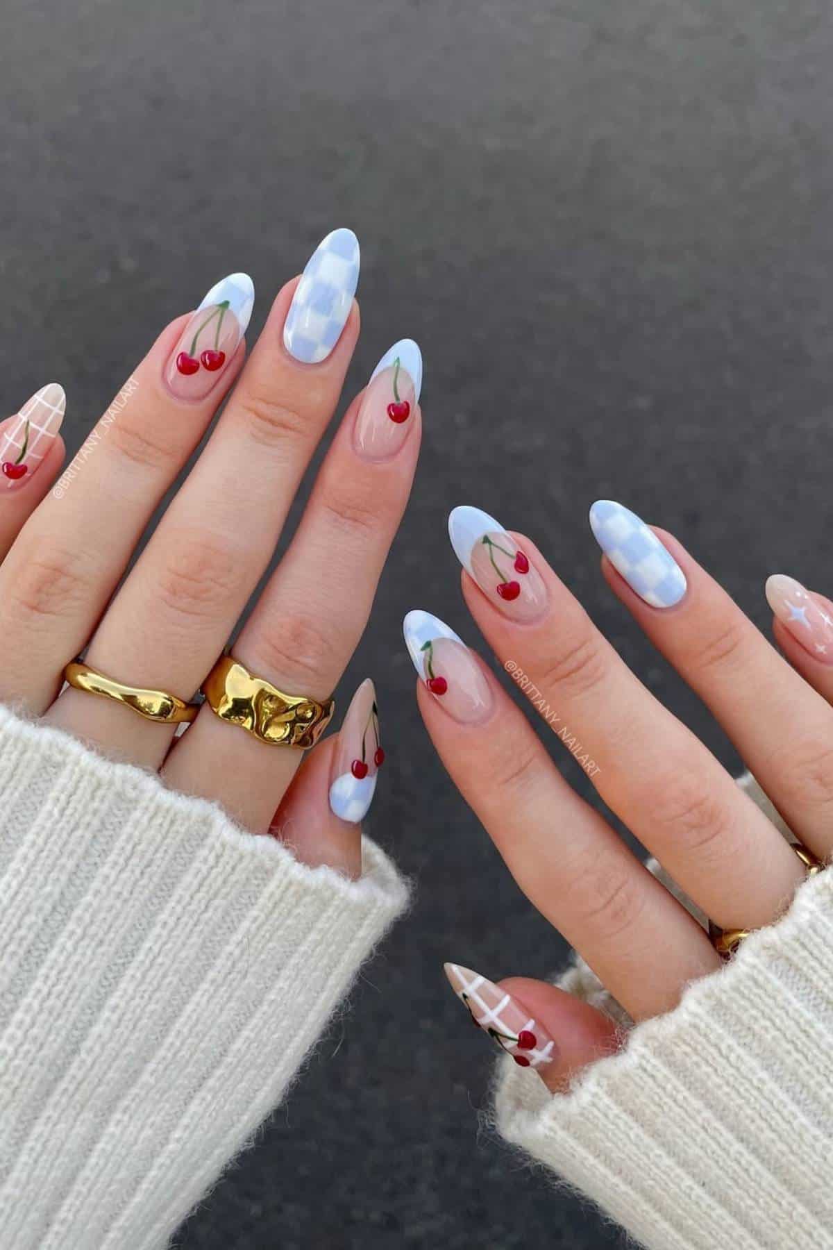 Hands with long almond-shaped nails featuring cherry and blue plaid nail art, wearing gold rings and a cream knit sweater, against a dark background.