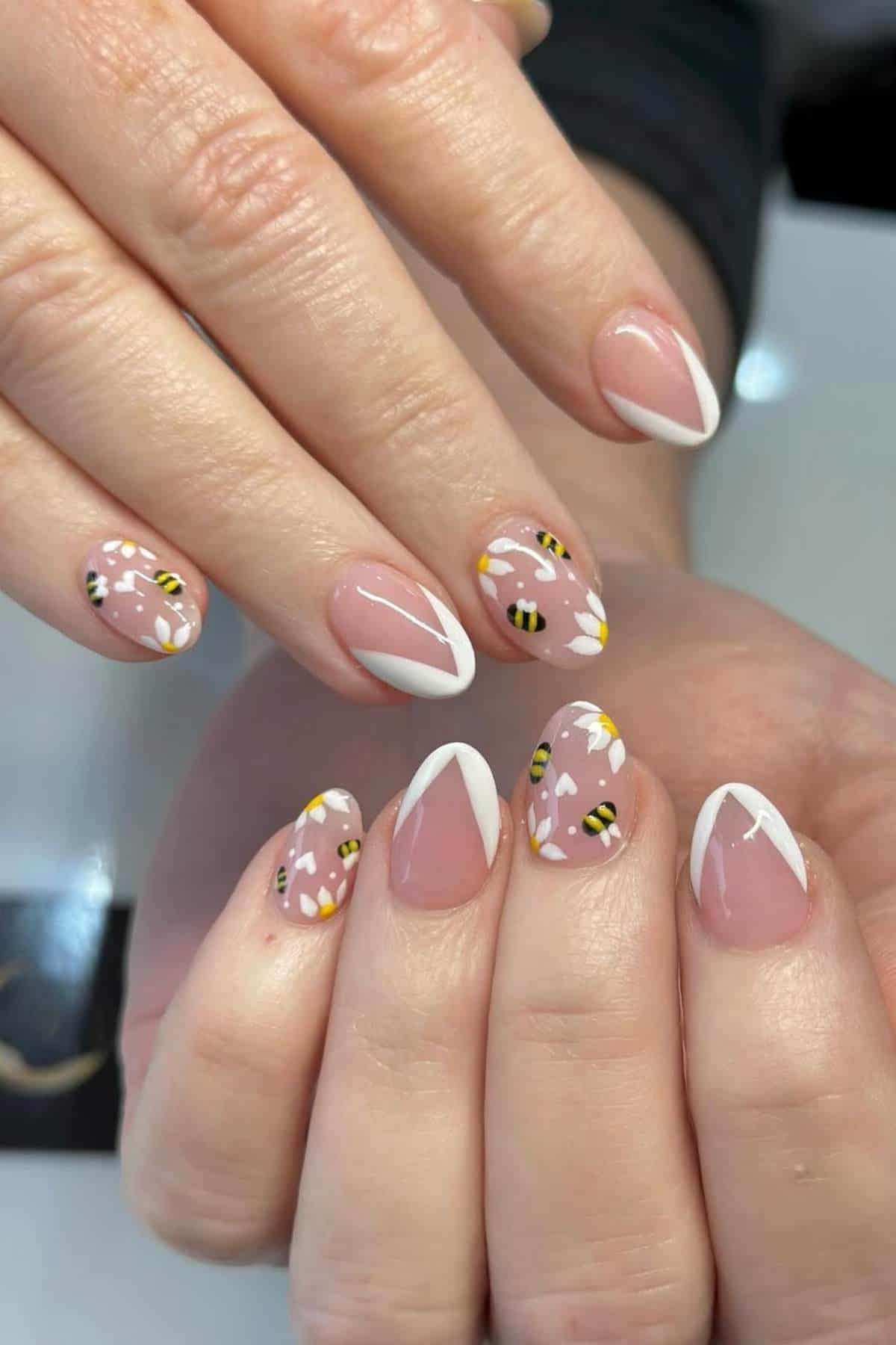 Hands with almond-shaped nails featuring French tips and bee-themed nail art with white and yellow floral designs on a nude pink base.