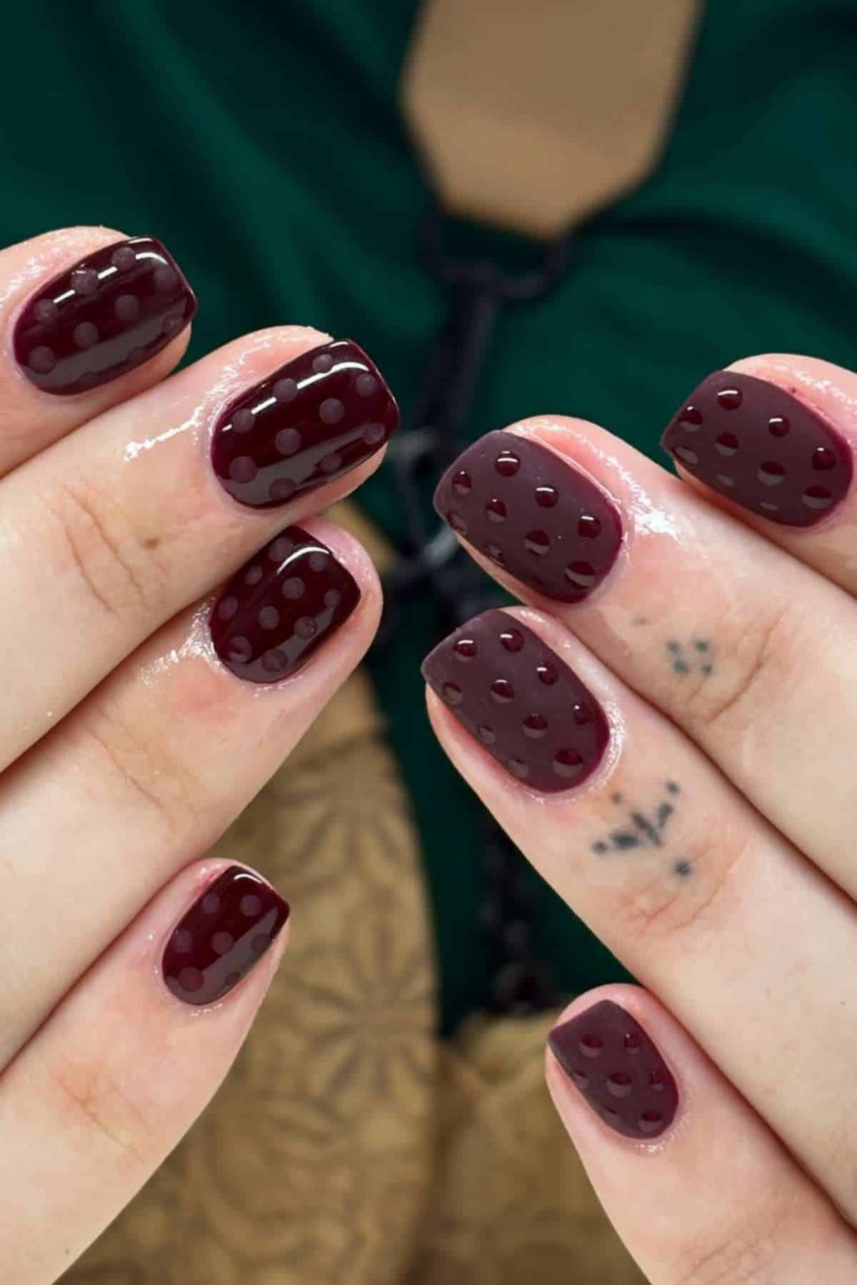 Close-up of hands with short, dark burgundy nails featuring a glossy finish and raised polka dot texture on each nail.