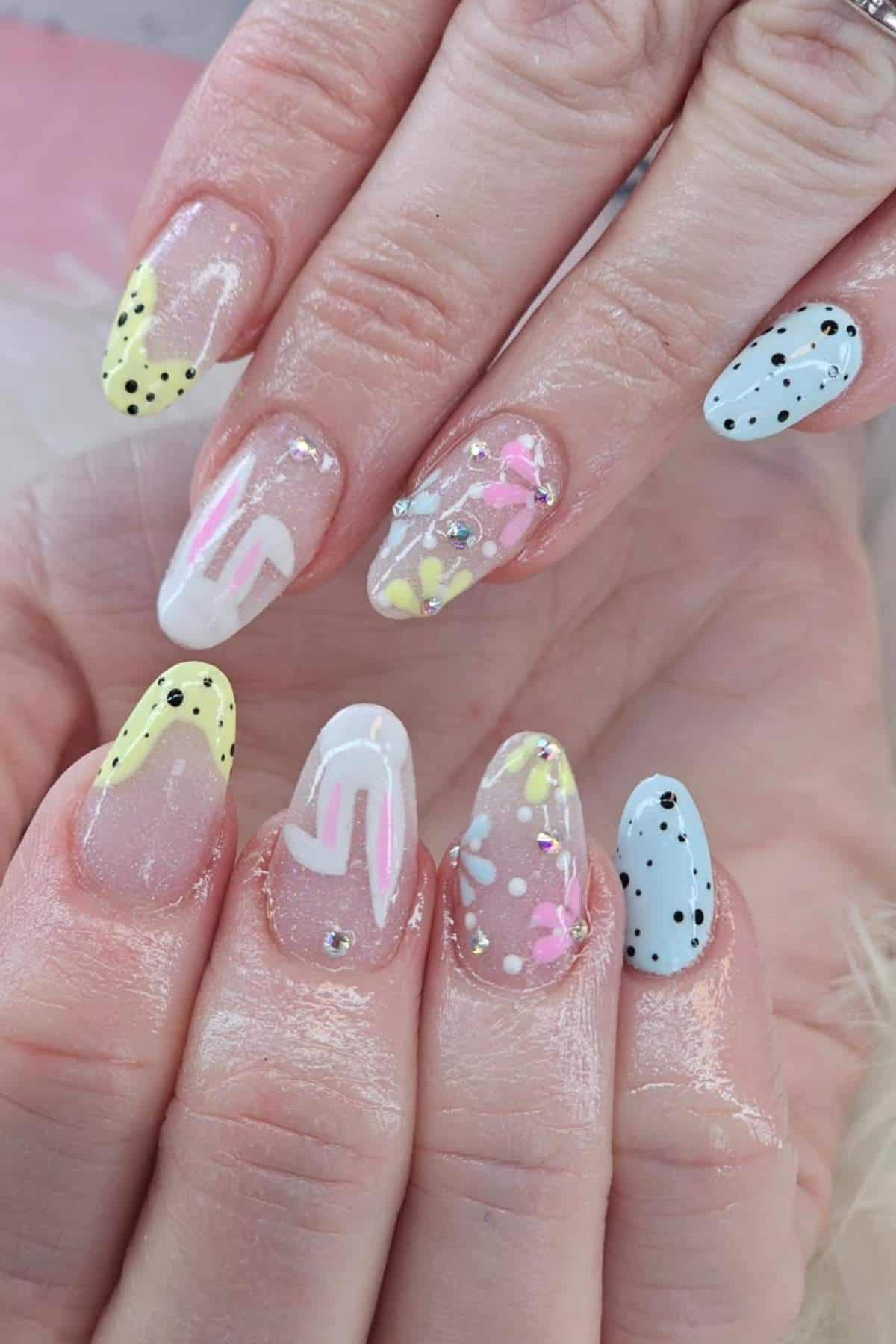 Close-up of hands with long, almond-shaped nails featuring pastel designs, including floral patterns, polka dots, geometric lines, and small rhinestone accents.