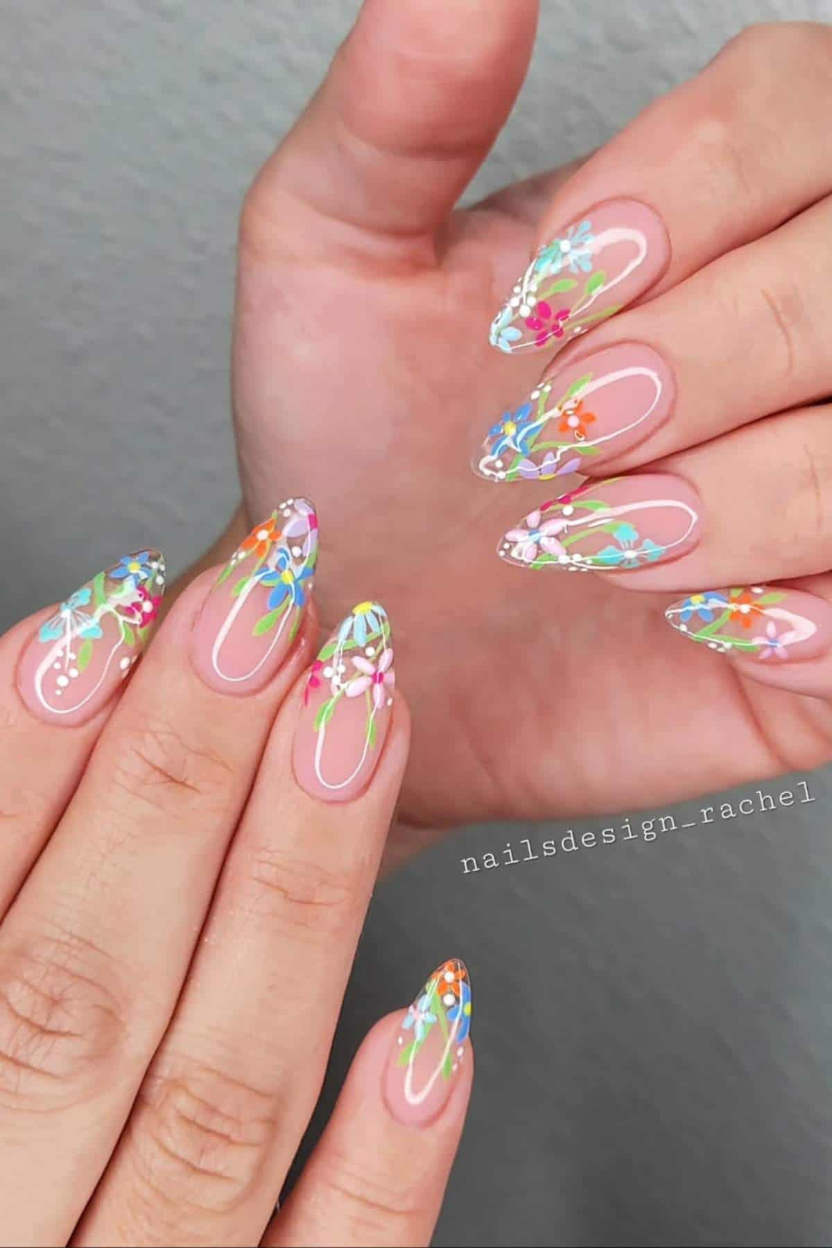 Hands with long almond-shaped nails featuring colorful floral designs and green lines on a nude base. The nails have a glossy finish.