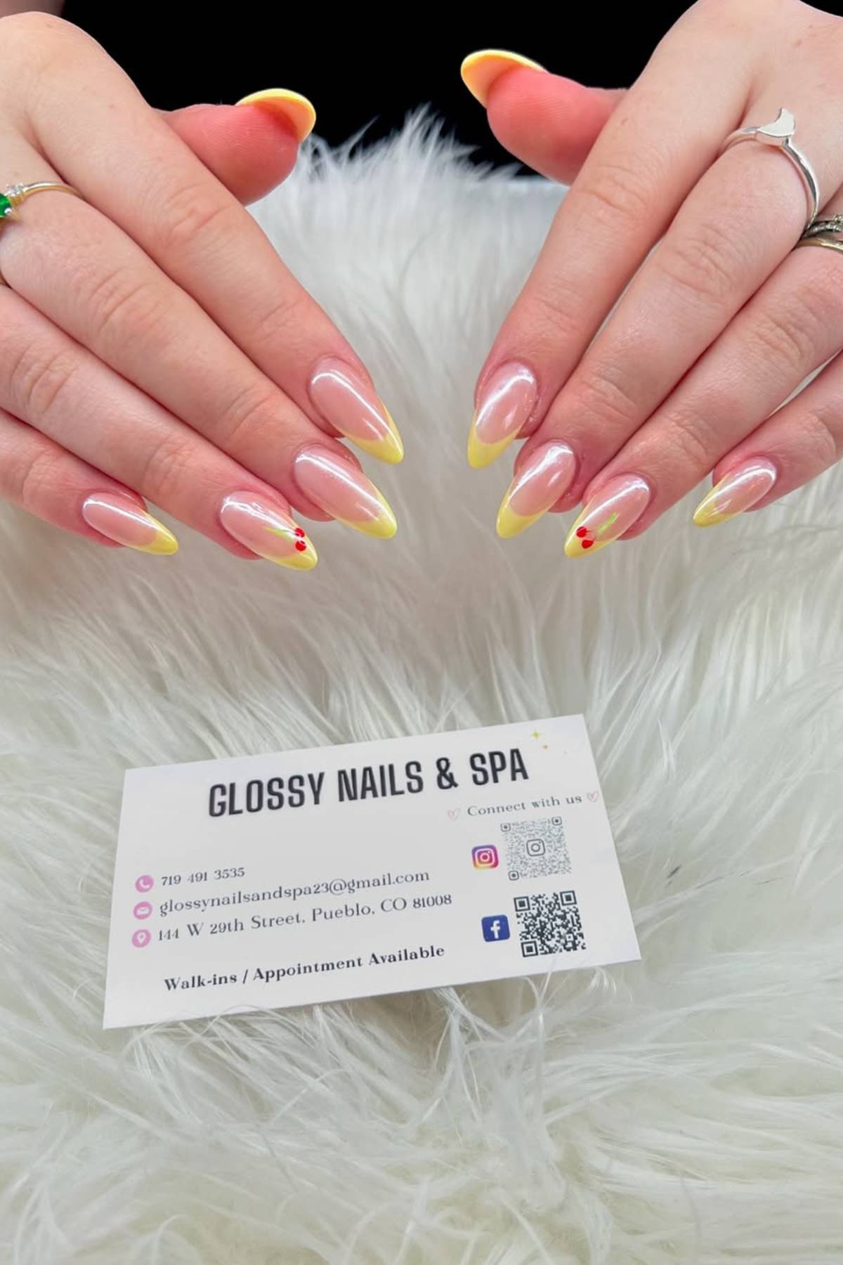Hands with long almond-shaped nails featuring yellow French tips and small red flower accents, displayed above a Glossy Nails & Spa business card on a white fur surface.