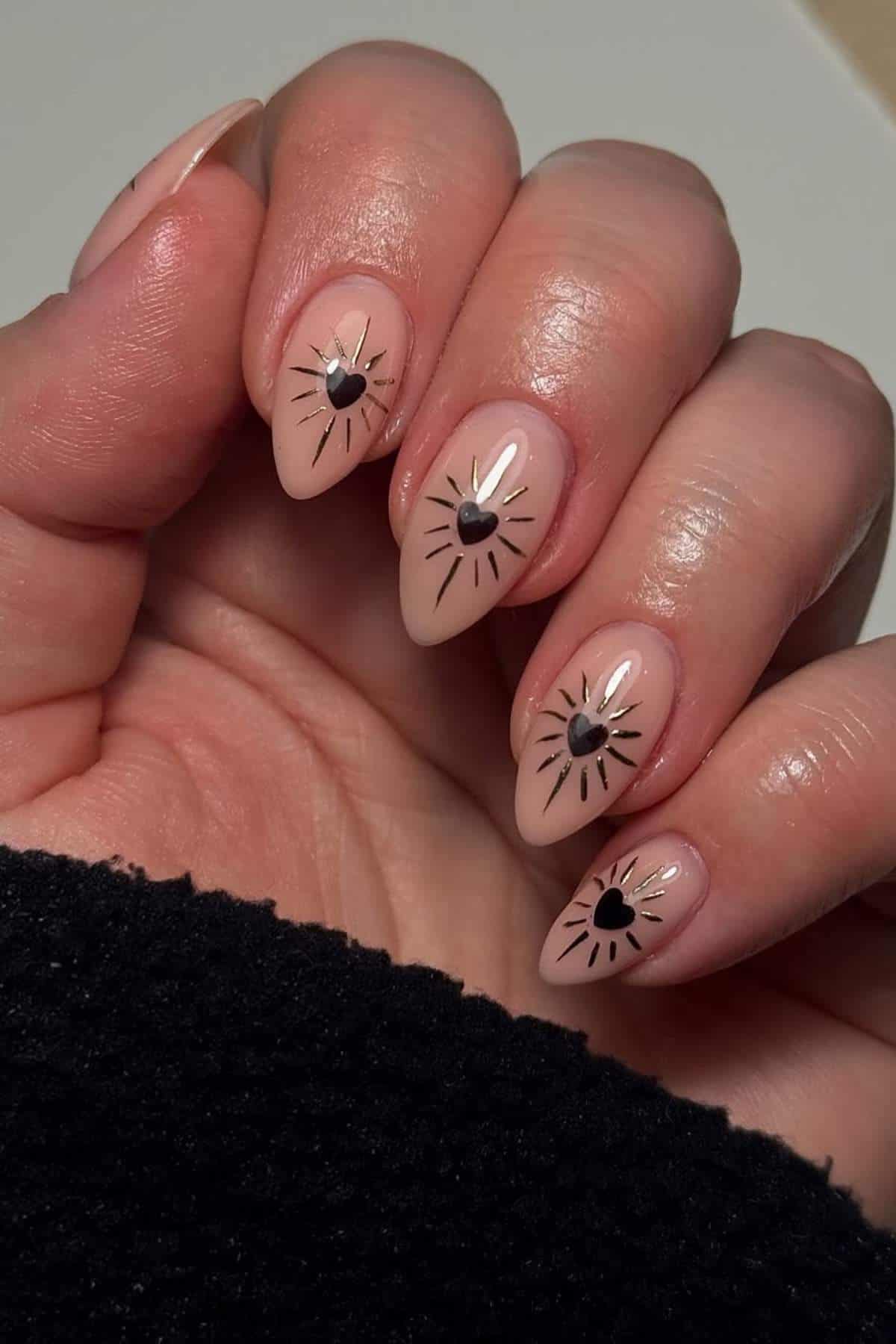 A hand with almond-shaped nails painted nude, each nail featuring a black heart in the center with black and gold radiating lines.