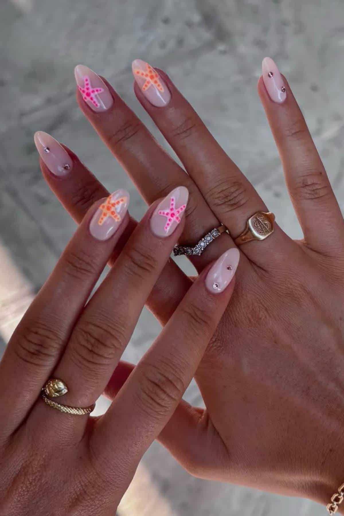 Hands with almond-shaped nails featuring nude polish, small rhinestones, and pink and orange starfish designs on several nails. The person is wearing gold rings.