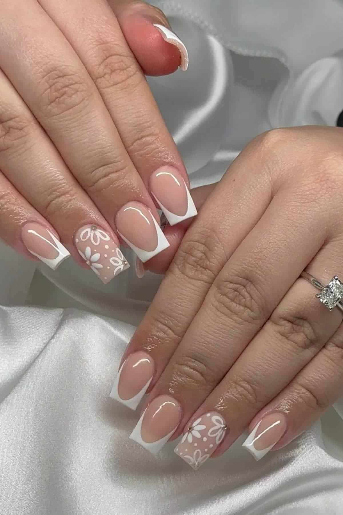 Hands with square-shaped French manicure on natural nails, featuring white floral designs on two accent nails, resting on white satin fabric. One hand wears a silver ring with a large gemstone.