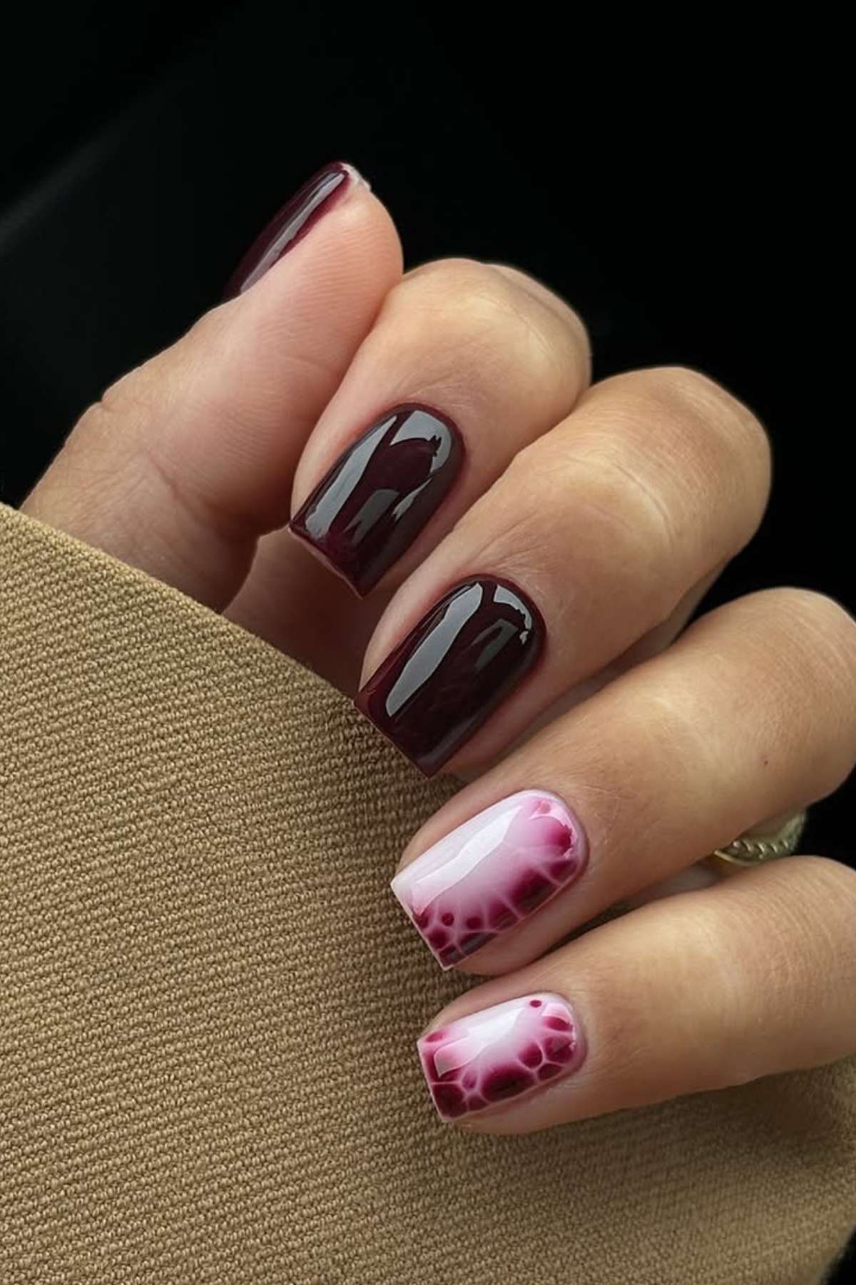 A hand with square-shaped nails; most are painted dark burgundy while two feature a pink and burgundy gradient with a reptile scale pattern.