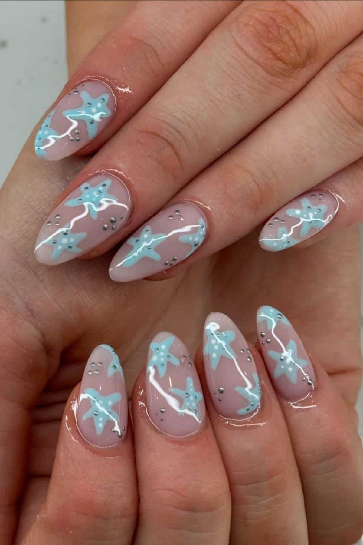 Close-up of hands with almond-shaped nails featuring a nude base, blue starfish designs, white wavy lines, and small silver dots as accents.