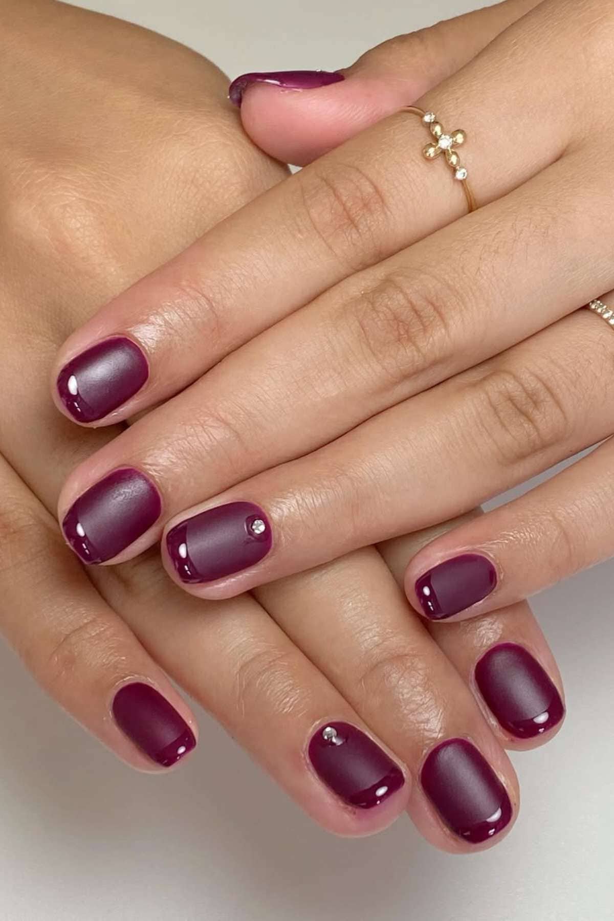 Hands with short, oval-shaped nails painted in glossy dark purple polish, each featuring a small silver rhinestone accent near the cuticle.