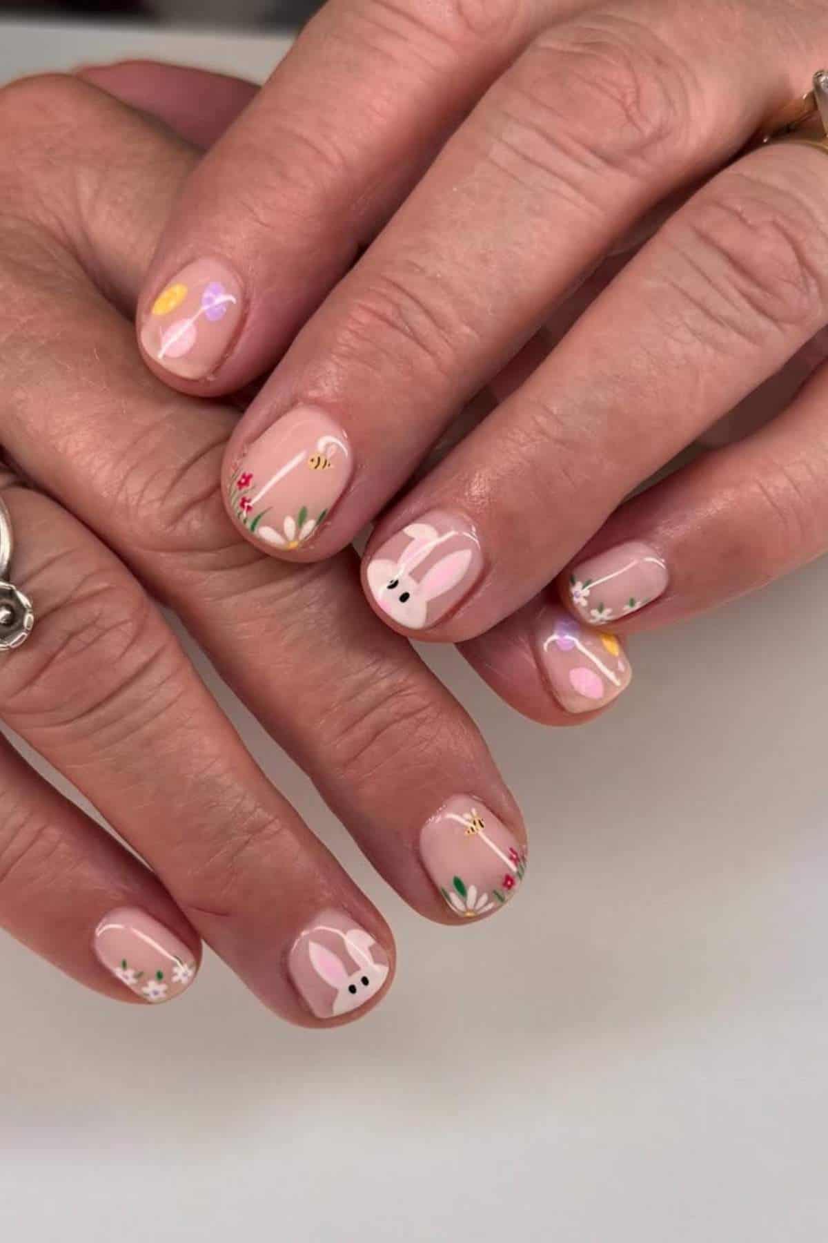 Hands with short nails featuring pastel Easter-themed nail art, including bunnies, flowers, and eggs on a light pink base.