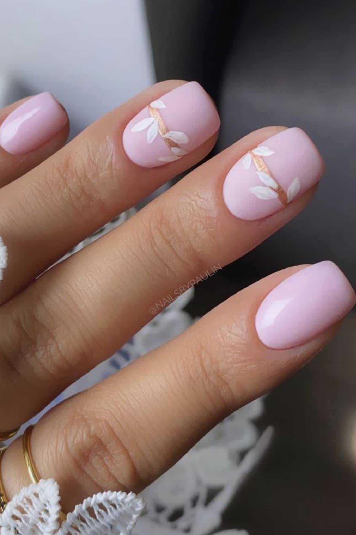 A hand with light pink manicured nails; two nails feature a delicate white leaf and gold branch design.