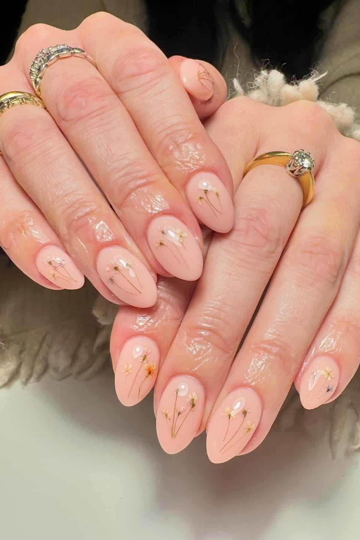 Hands with almond-shaped, pale pink manicured nails featuring delicate gold floral designs and small pearl accents, with rings on several fingers.
