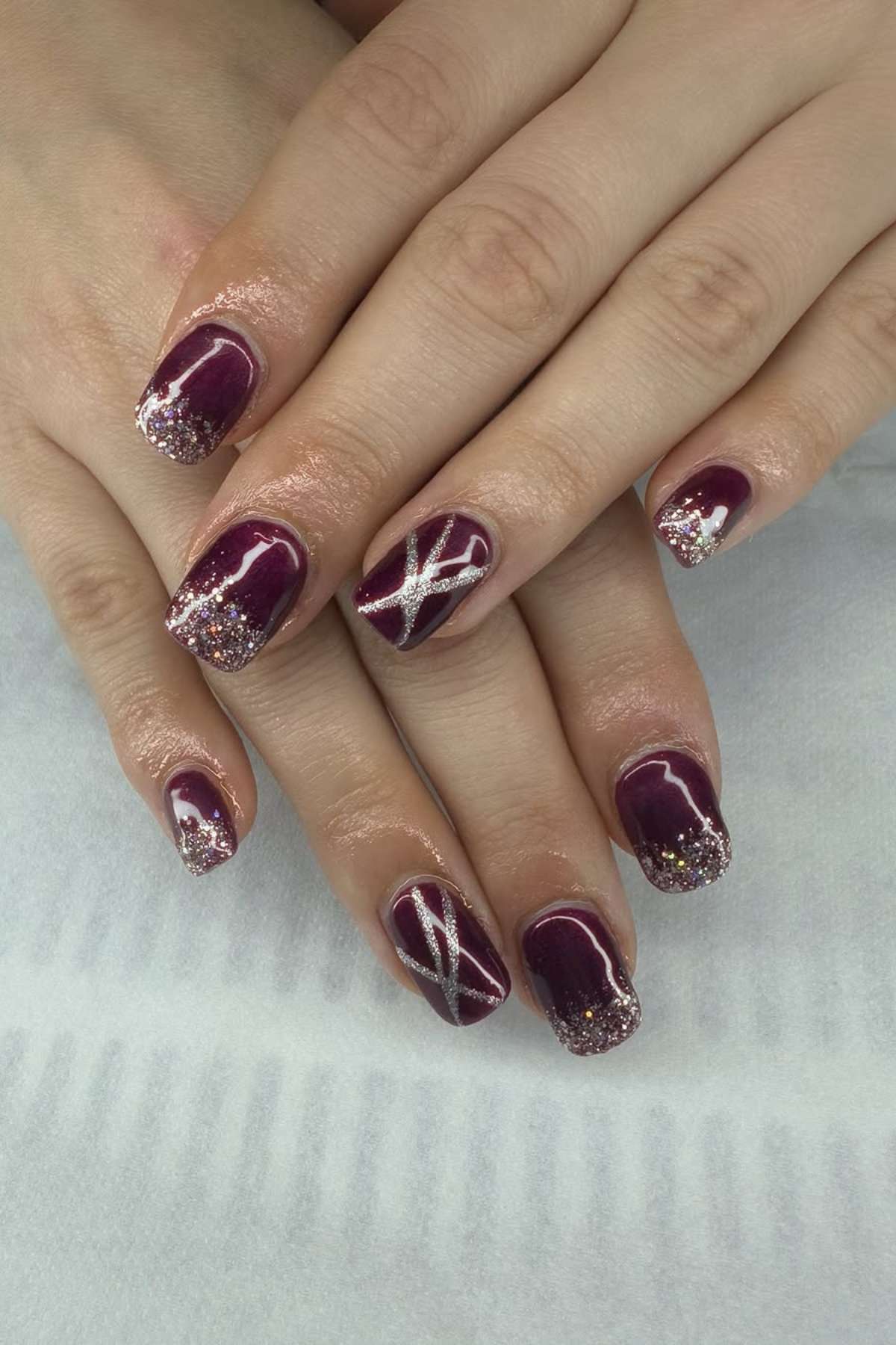 Hands with maroon nail polish featuring silver glitter tips and accent nails with silver crisscross designs, resting on a white surface.