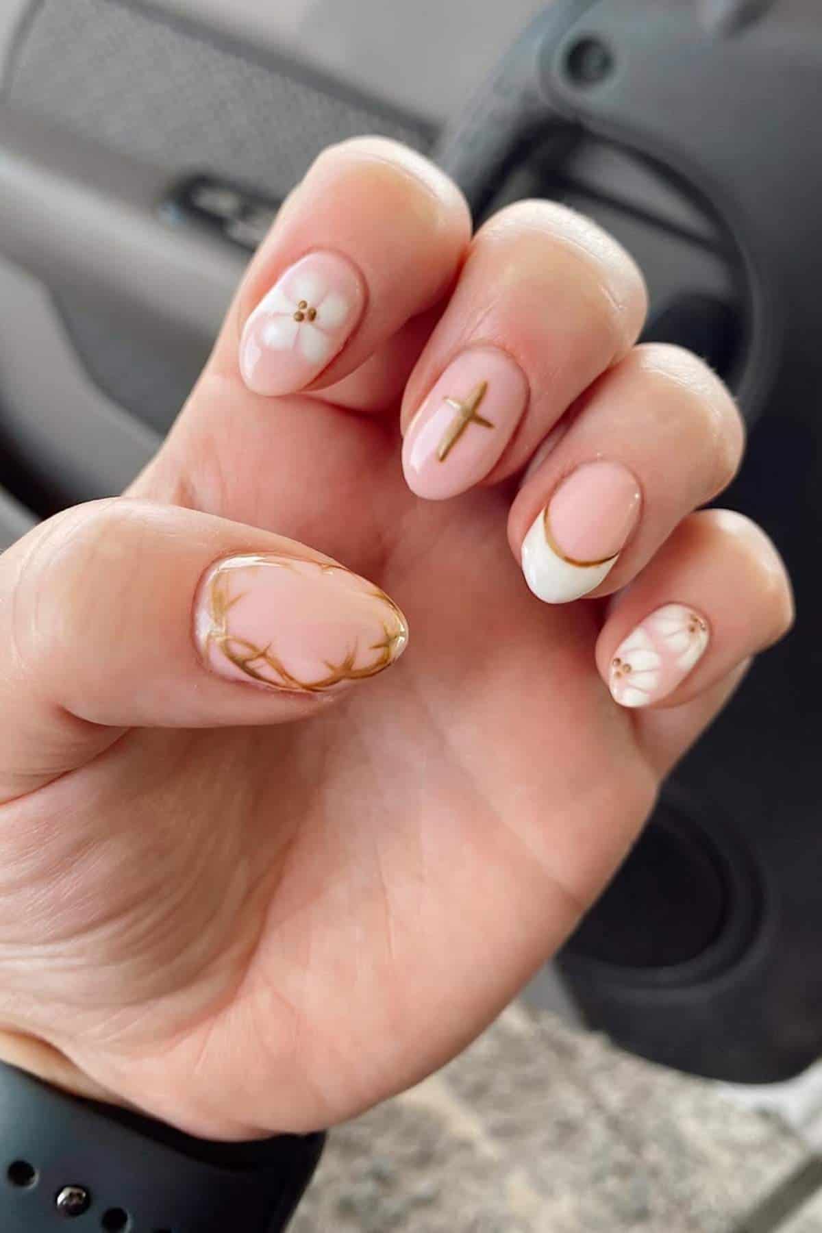A hand with long, almond-shaped nails painted in light pink with gold and white designs, including a cross, crown of thorns, and pearl-like accents.