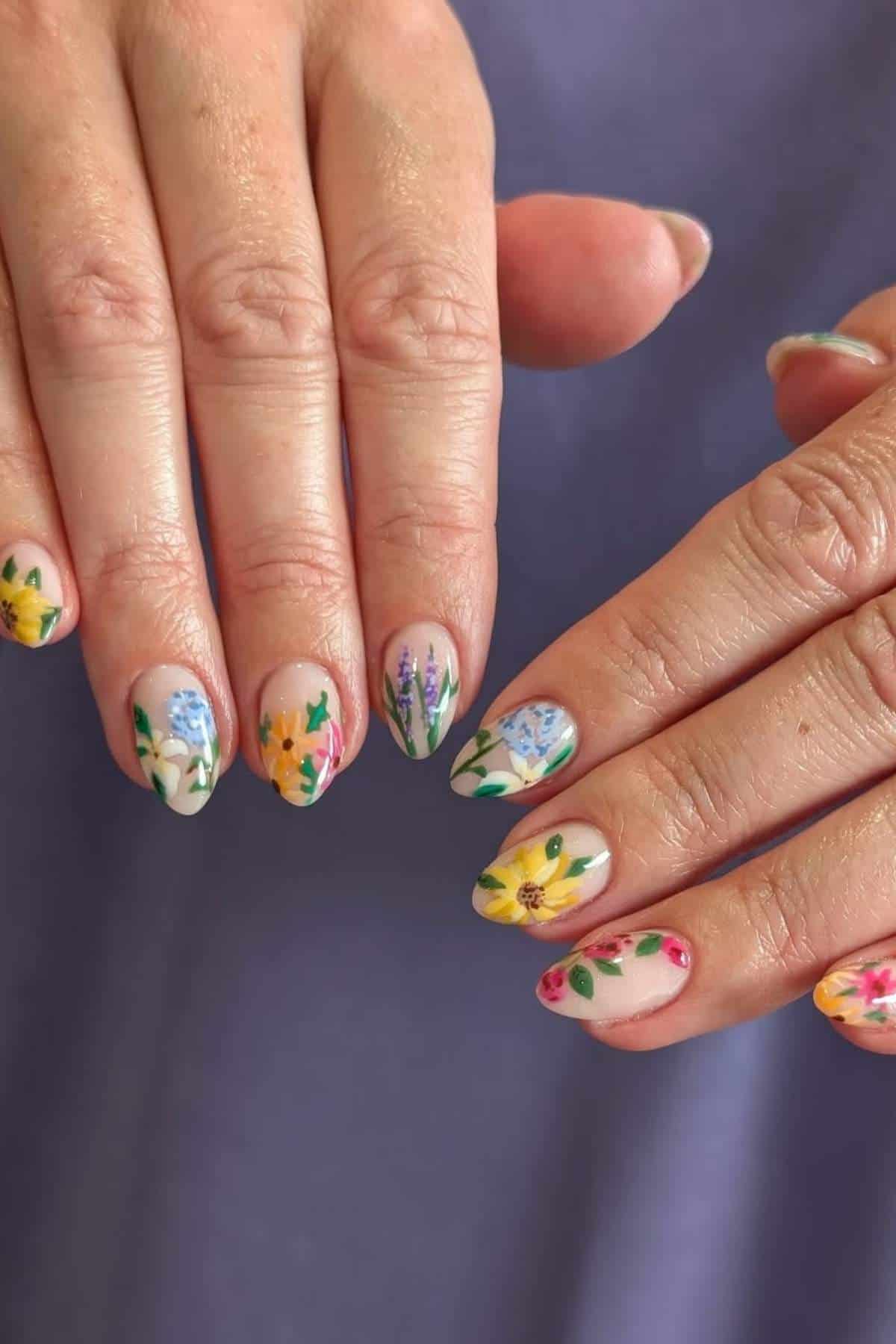 Close-up of hands with almond-shaped nails painted with colorful floral designs against a purple background.