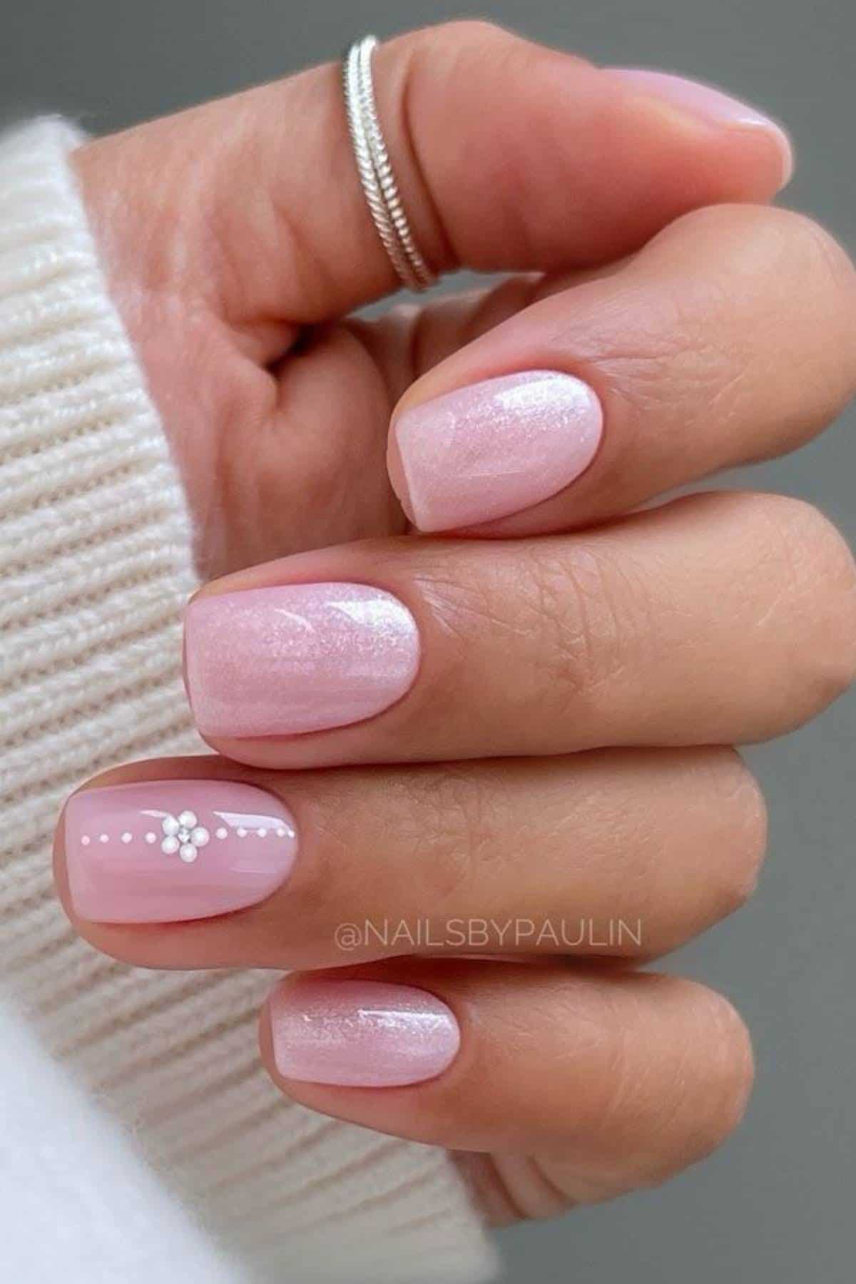 A hand with short, square-shaped nails painted light pink with a subtle shimmer; one nail features a small white flower and dotted design.