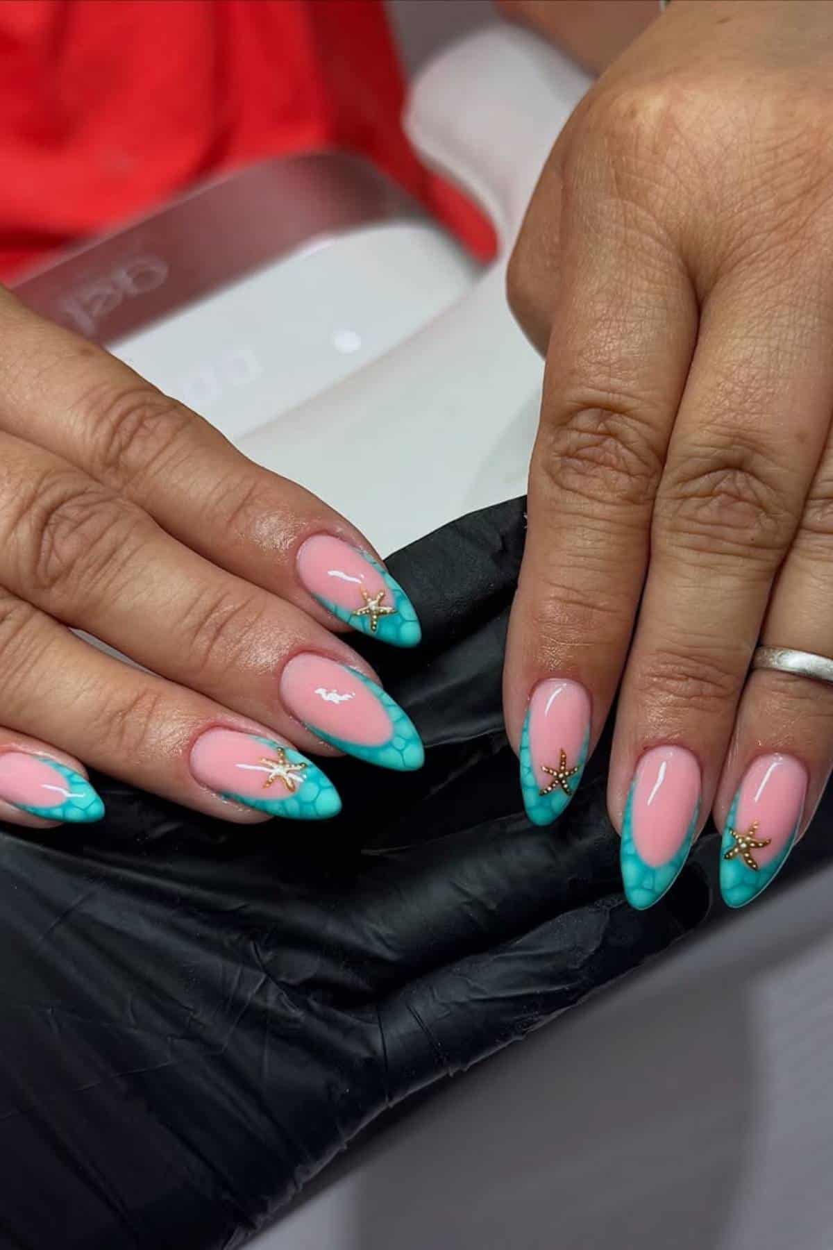 Hands with long almond-shaped nails featuring pink polish, turquoise tips with a scale pattern, and small gold starfish decorations.