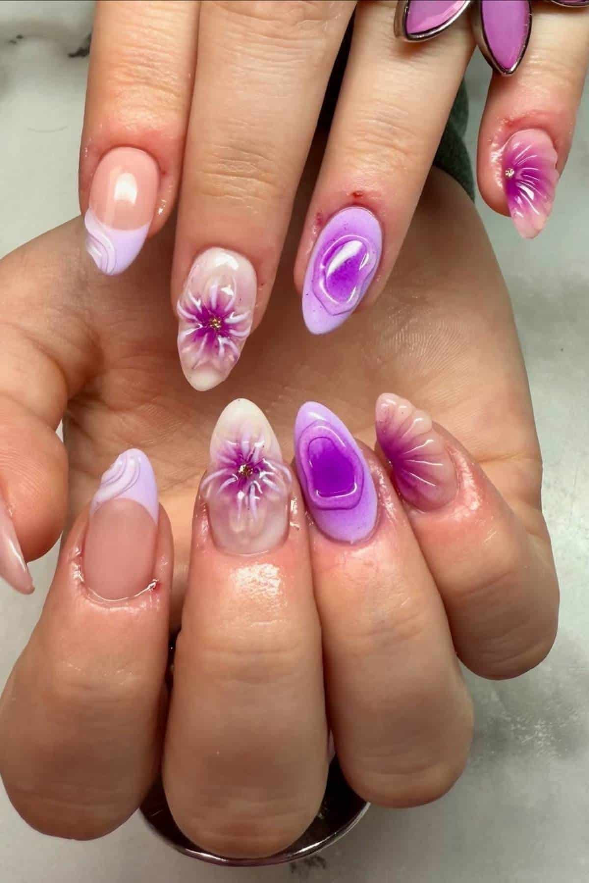 Close-up of hands with manicured nails featuring purple and pink floral designs, abstract patterns, and glossy finishes.