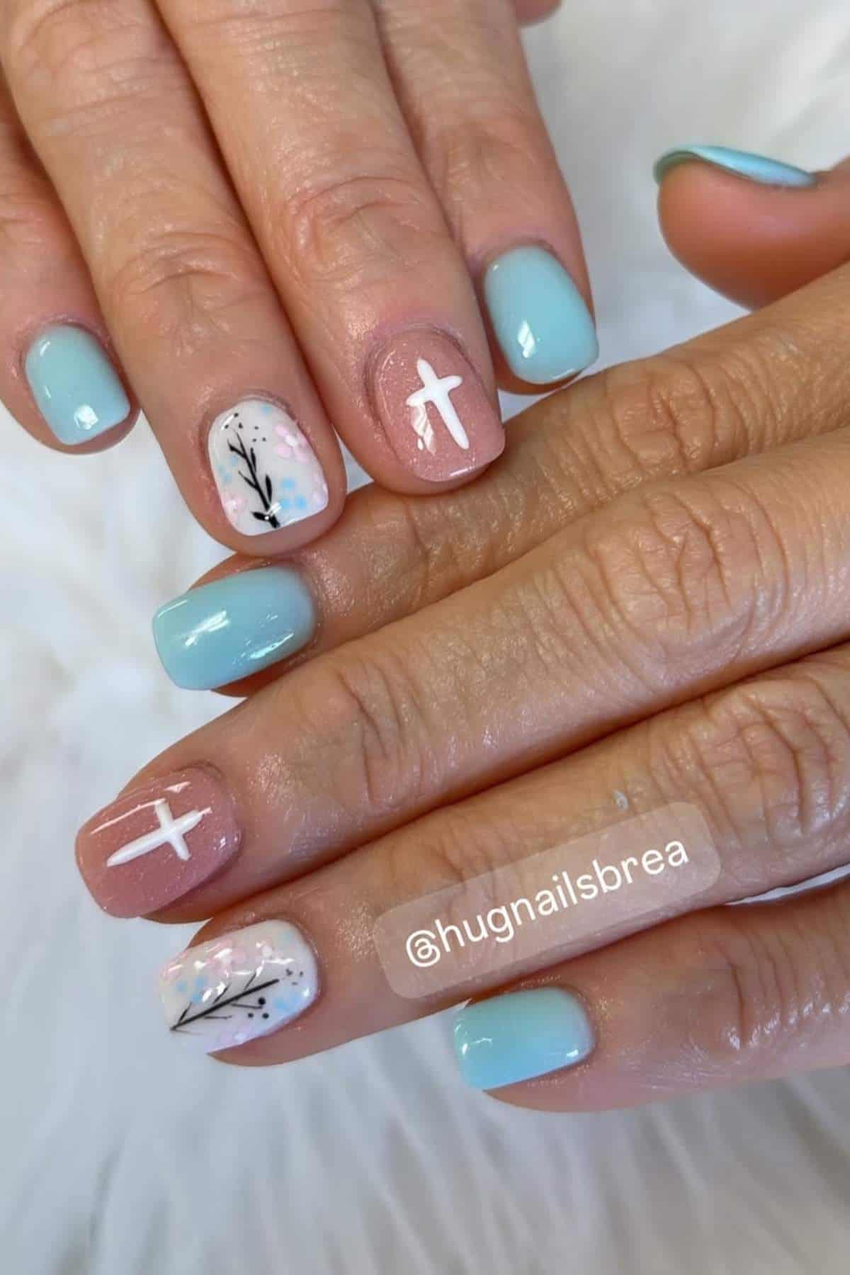 Hands with short square nails painted in pastel blue and nude, featuring white crosses and black branch designs as accent nail art.