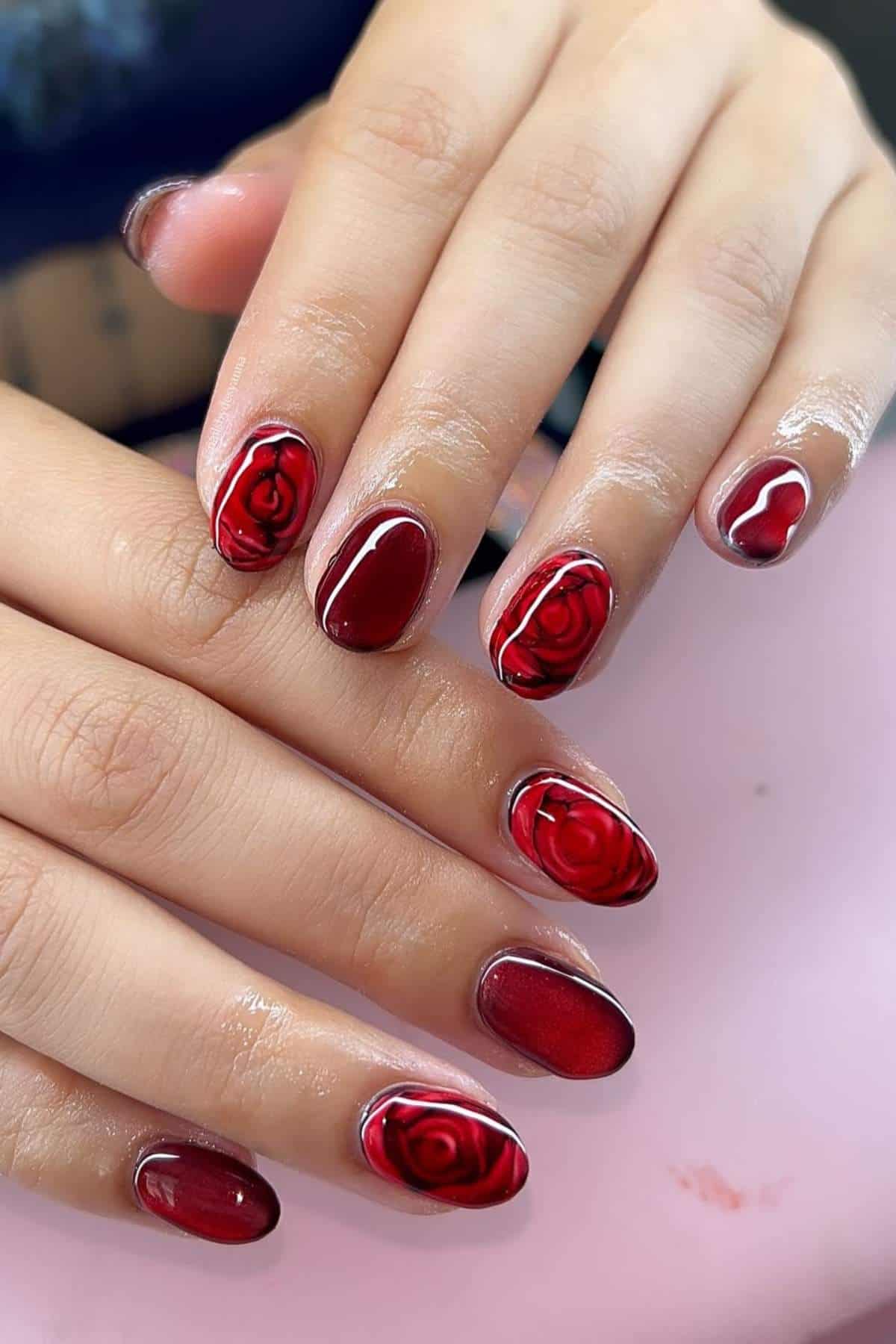 A hand with glossy red manicure featuring rose designs on some nails, displayed against a soft pink background.