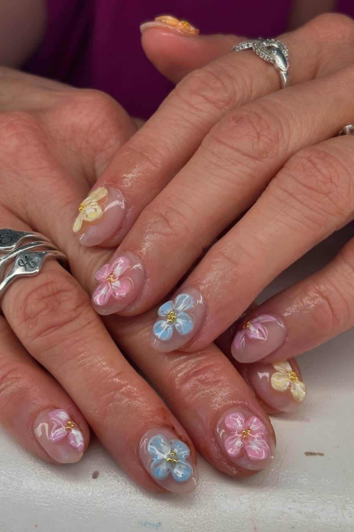 Close-up of hands with pastel-colored 3D flower nail art on natural, short, oval-shaped nails. The person is wearing silver rings.