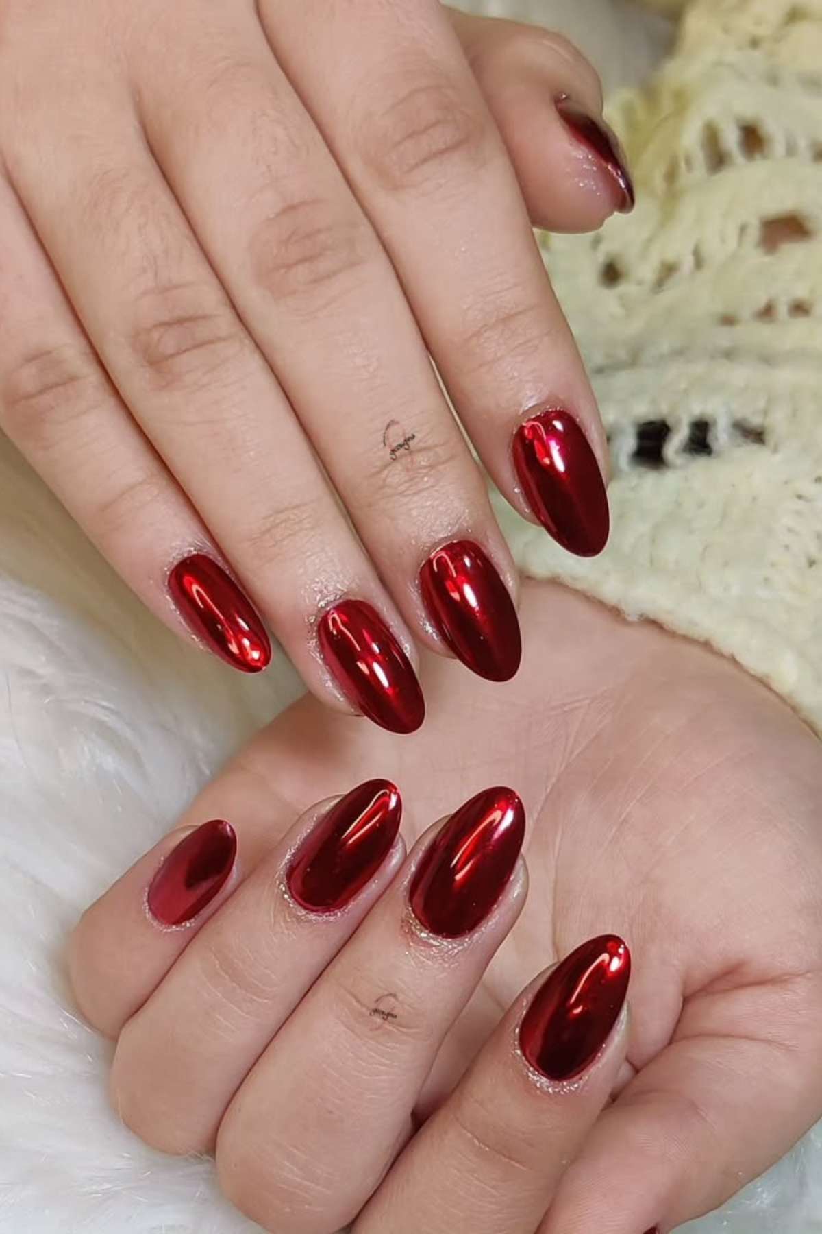 Close-up of hands with almond-shaped nails painted in glossy red polish, resting on a light surface.