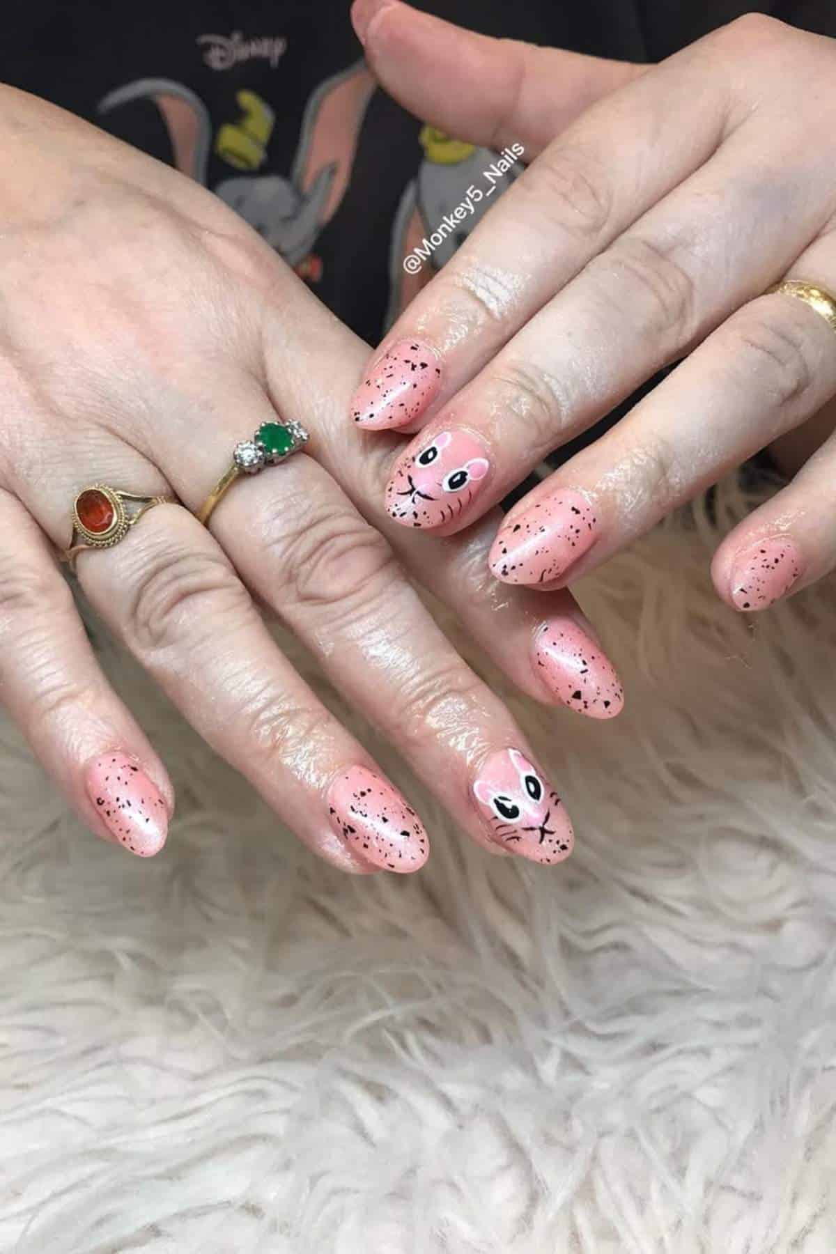 Two hands with pink, speckled nail art; some nails feature cartoon bunny faces. The person wears rings and rests their hands on a white furry surface.