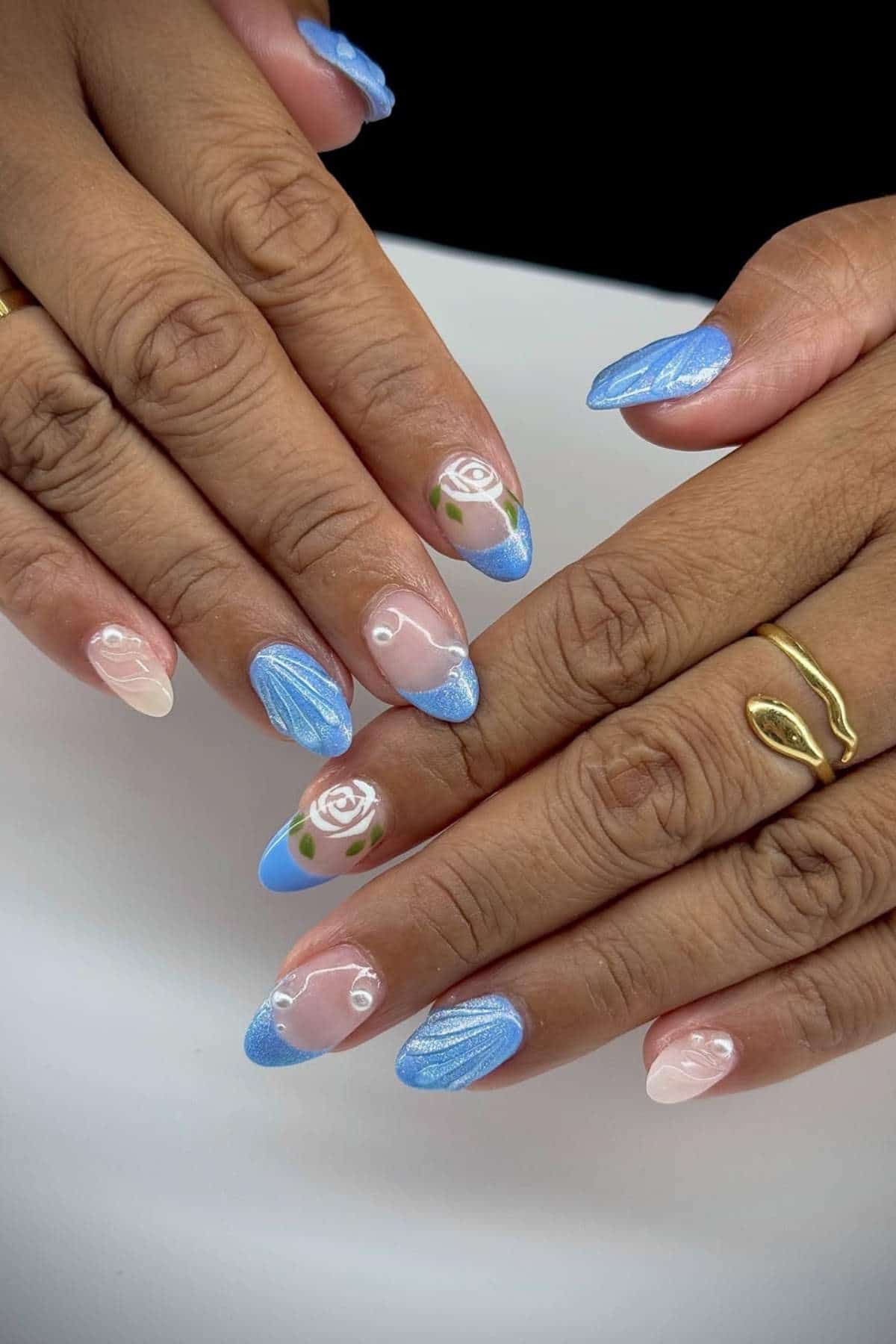 Hands with almond-shaped nails featuring blue glitter French tips, white rose designs on two nails, and subtle white swirl accents on a nude base.