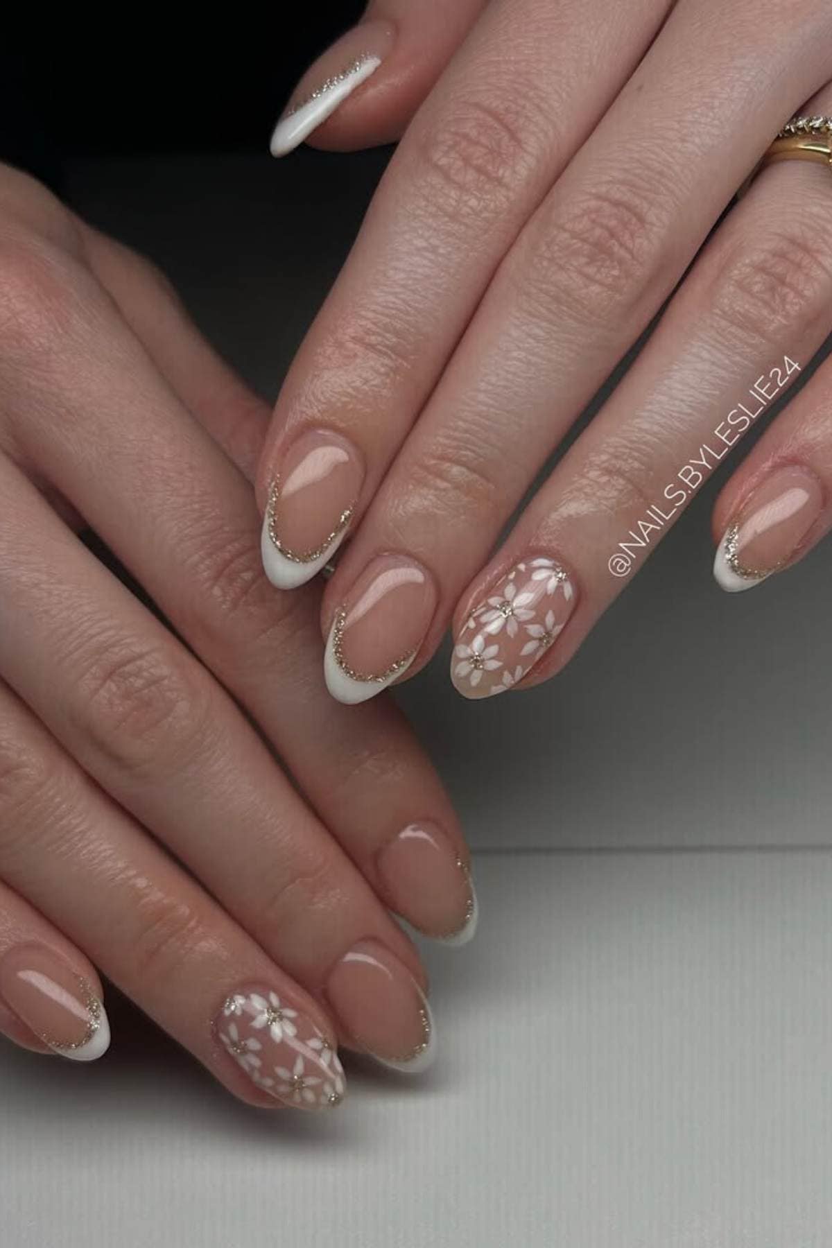 Hands with almond-shaped nails featuring a French tip design, gold accents, and white floral patterns on two accent nails.