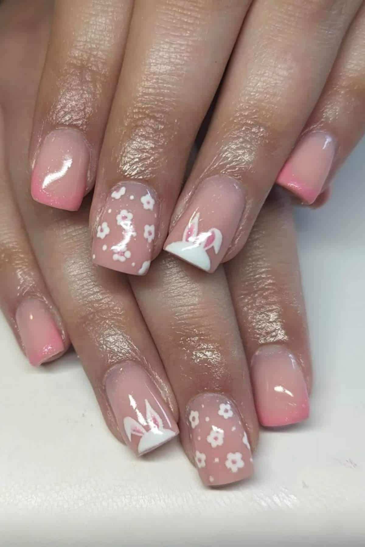 Close-up of hands with square-shaped nails featuring pink ombré, white floral patterns, and white abstract line art designs on a few accent nails.