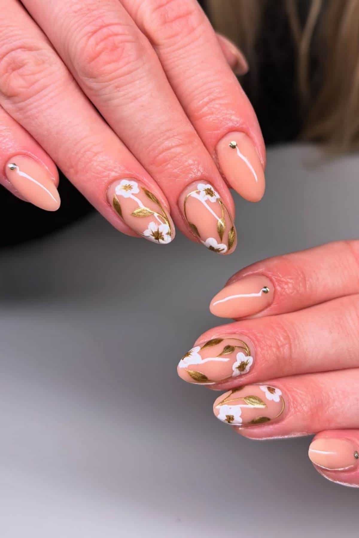 Hands with almond-shaped nude nails, decorated with white flower designs, green leaves, and small rhinestones on a few nails.