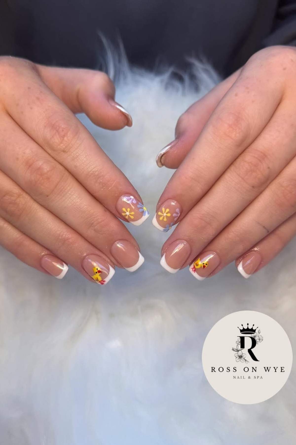 A pair of hands with French manicured nails featuring yellow and red floral designs on some fingers, displayed on a fluffy white surface. “Ross On Wye Nail & Spa” logo in the corner.