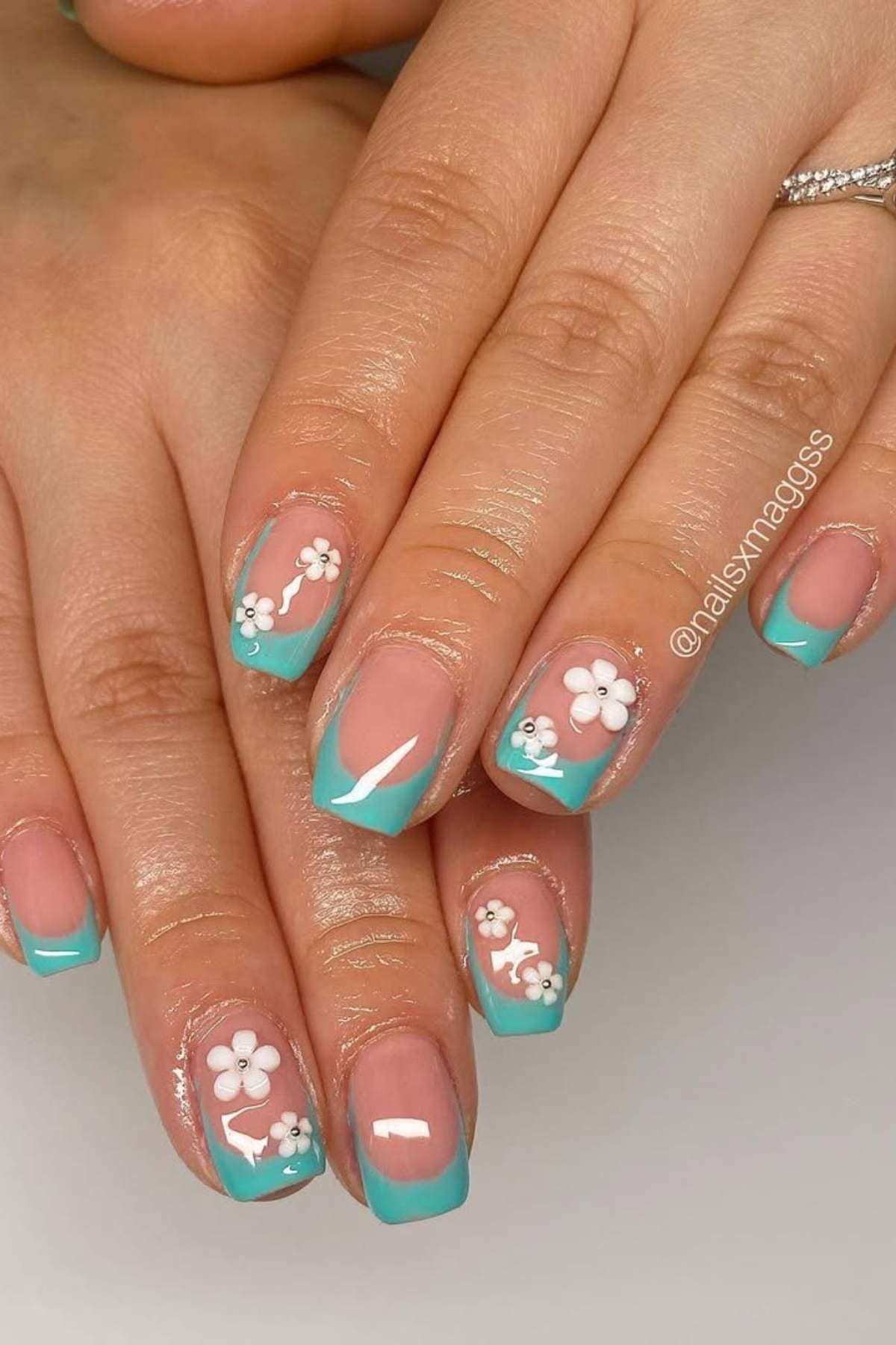 Close-up of hands with turquoise French tip nails featuring white and pink floral designs on a nude base.