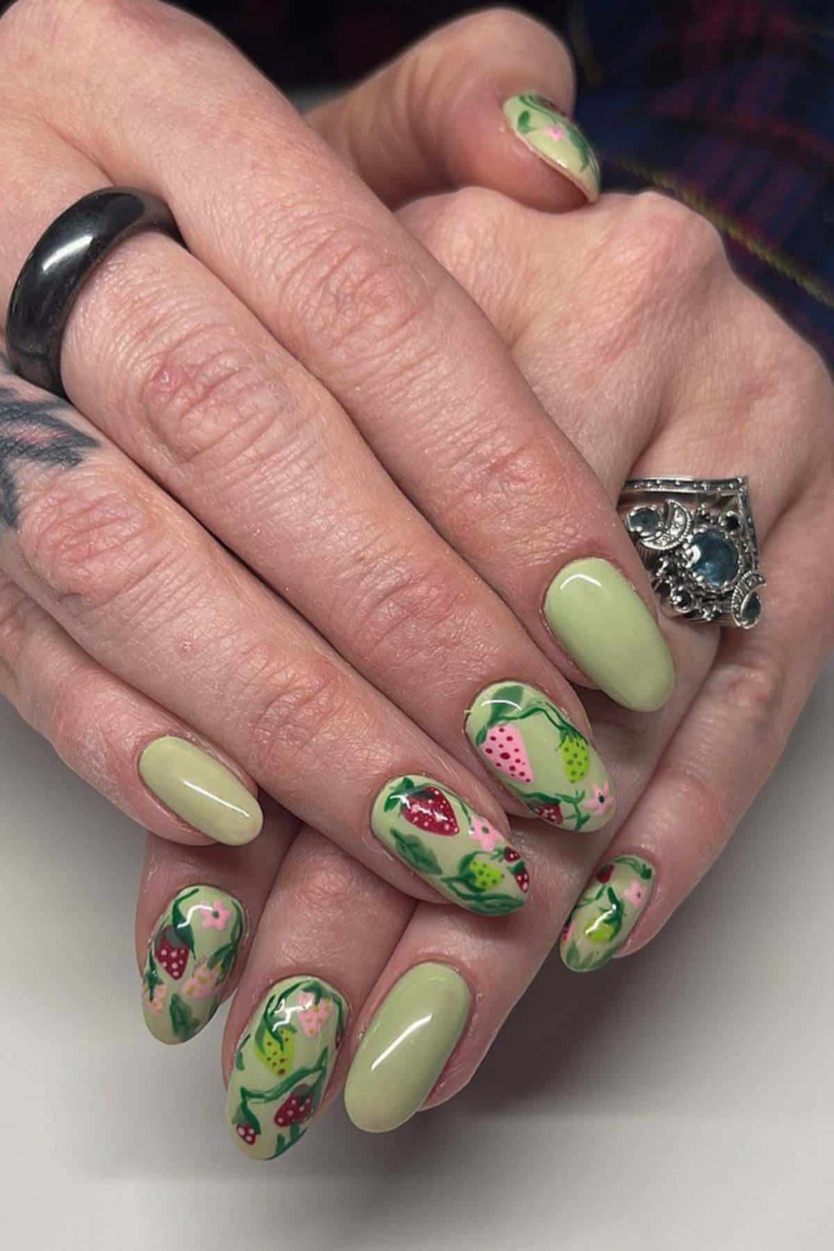 Hands with light green manicured nails, some featuring detailed fruit designs including strawberries and kiwis. The person wears a silver ring and a black ring.