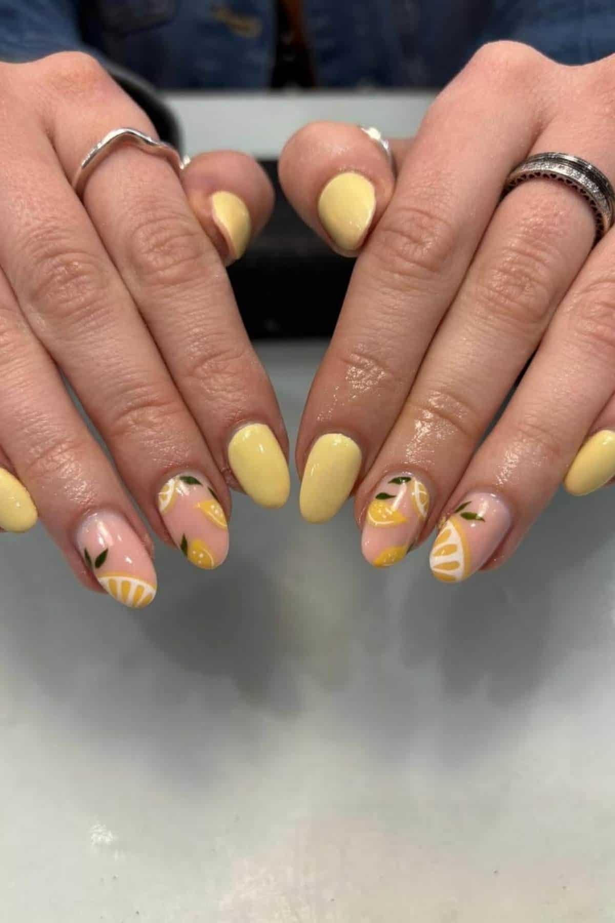 Hands with almond-shaped nails painted yellow, featuring lemon-themed designs and green leaf accents on some nails.