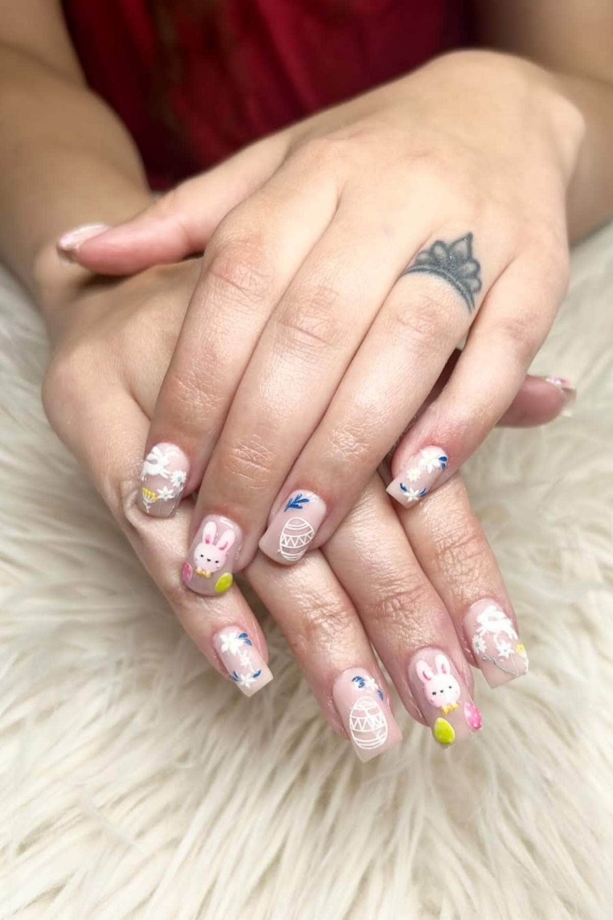 Hands with light pink, Easter-themed nail art featuring bunnies, eggs, flowers, and pastel decorations, resting on a white textured surface. A blue tattoo is visible on one finger.