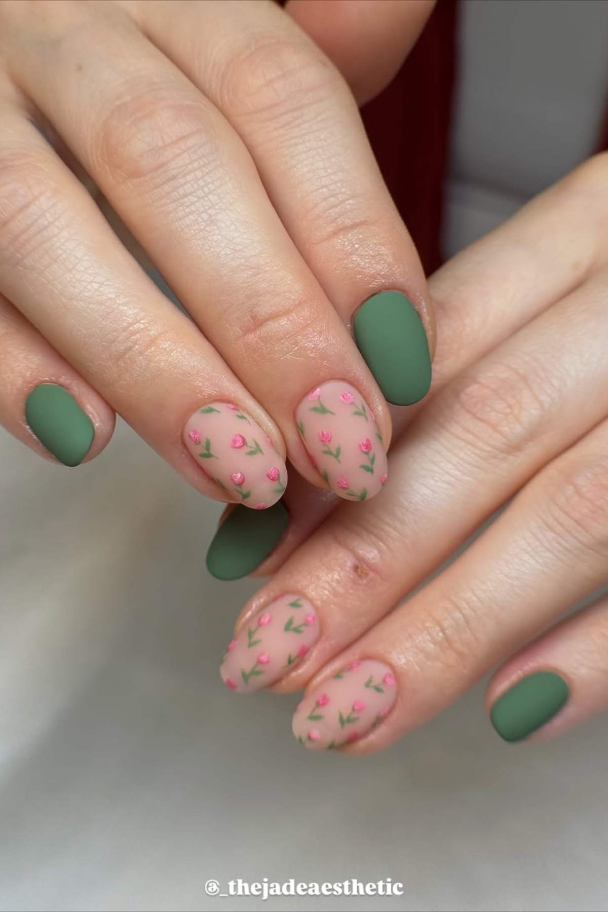 Hands with matte green and nude nails; nude nails feature small pink floral designs.