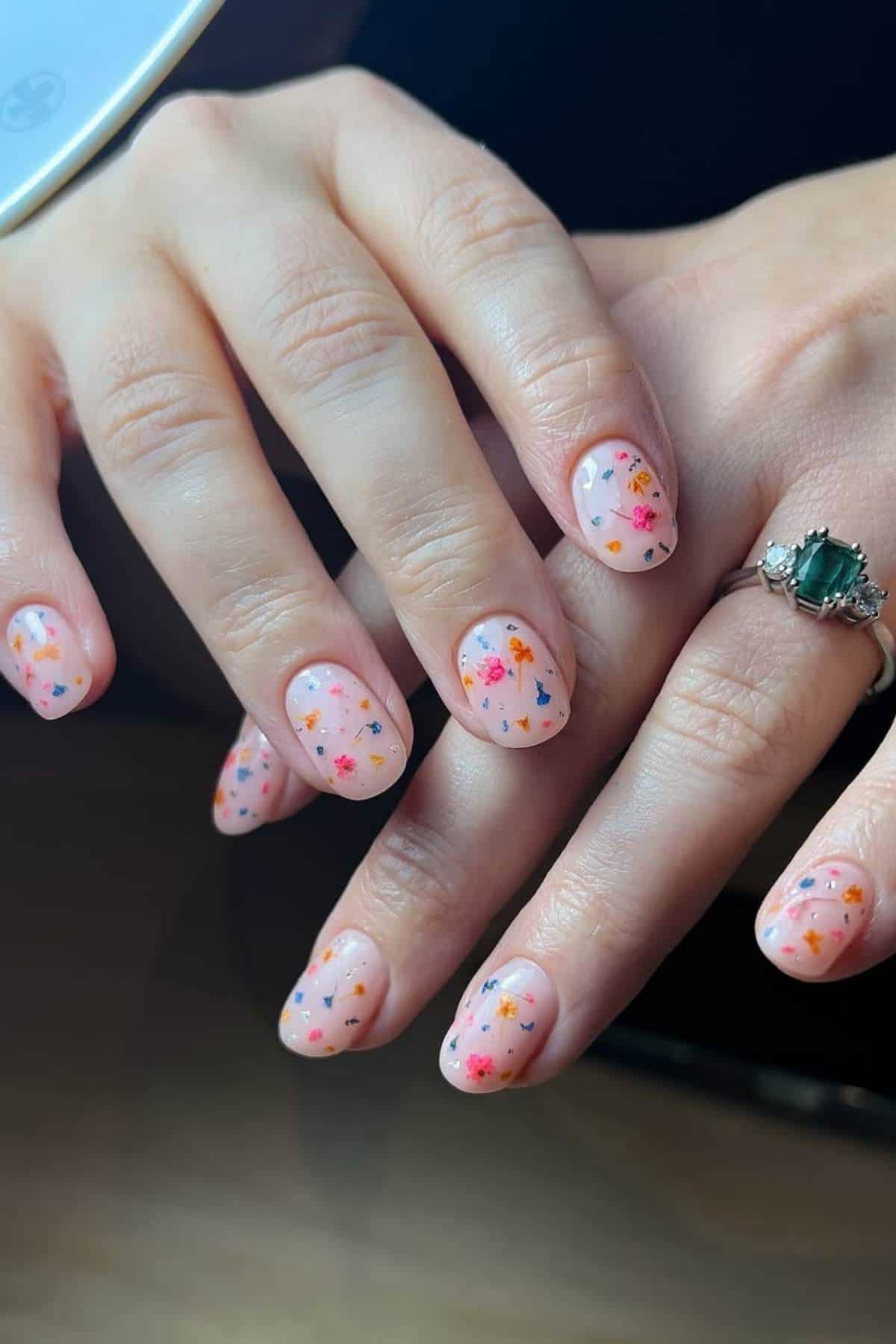 Close-up of hands with short, oval nails painted light pink and decorated with colorful small star and heart-shaped confetti; a green gemstone ring is worn on one finger.