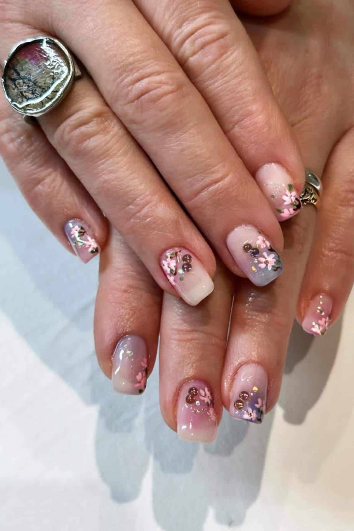 Hands with manicured nails featuring pink, purple, and white floral nail art with small gold accents. One hand wears a large silver ring with a marbled design.