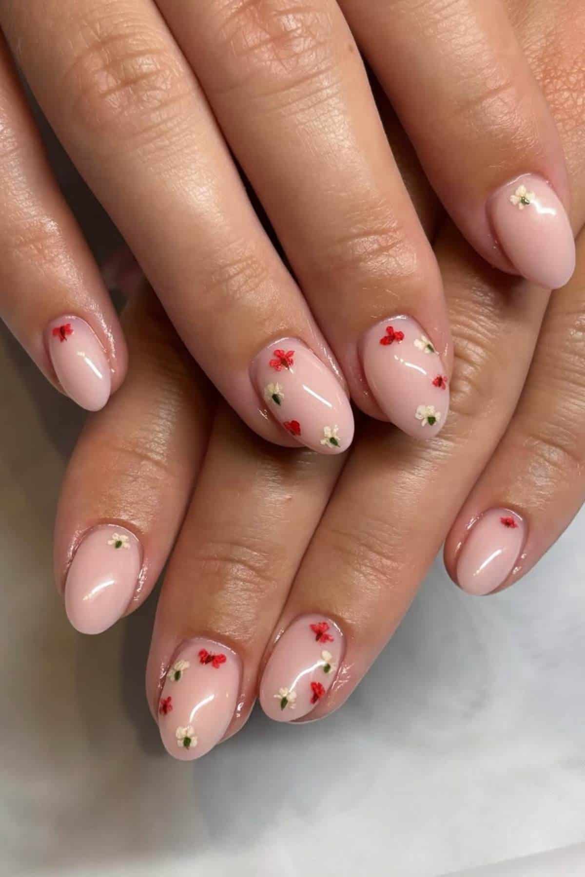 Hands with short, oval-shaped nails painted light pink, featuring small red and white flower designs on each nail.