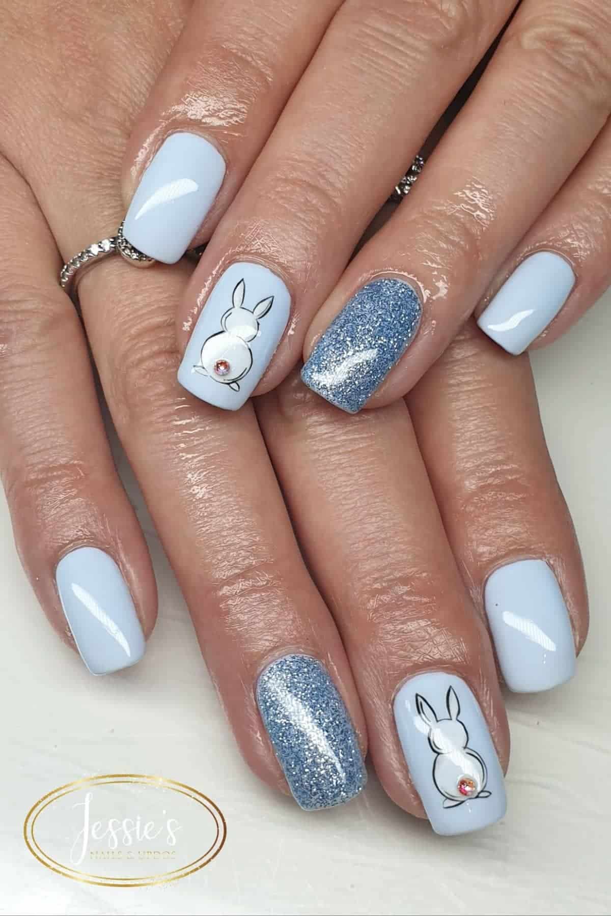 Hands with light blue manicured nails; some nails have glittery blue polish and others have white bunny designs with small rhinestones.