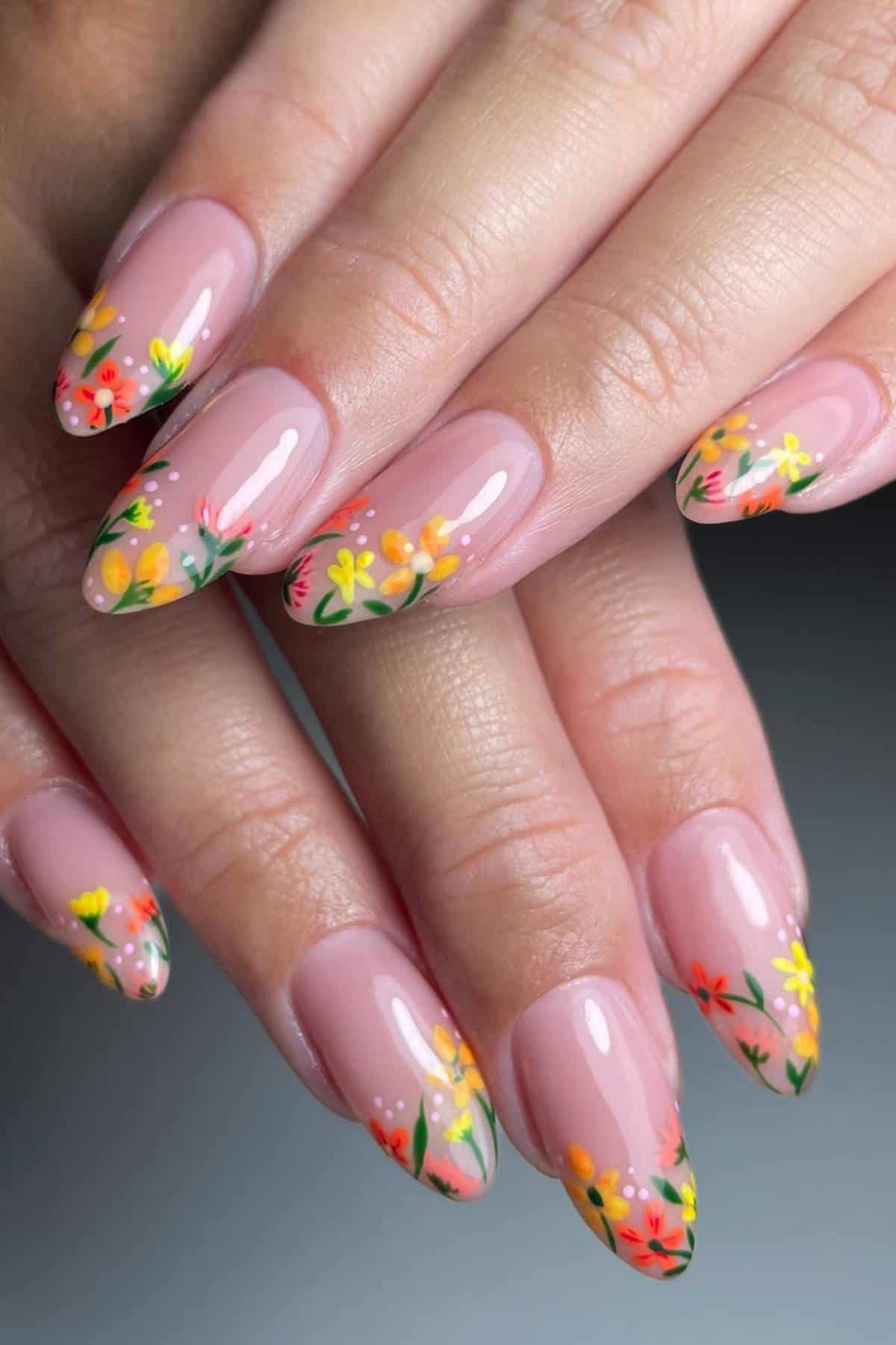 Close-up of hands with almond-shaped nails featuring a glossy nude base and colorful floral nail art in yellow, orange, and green.