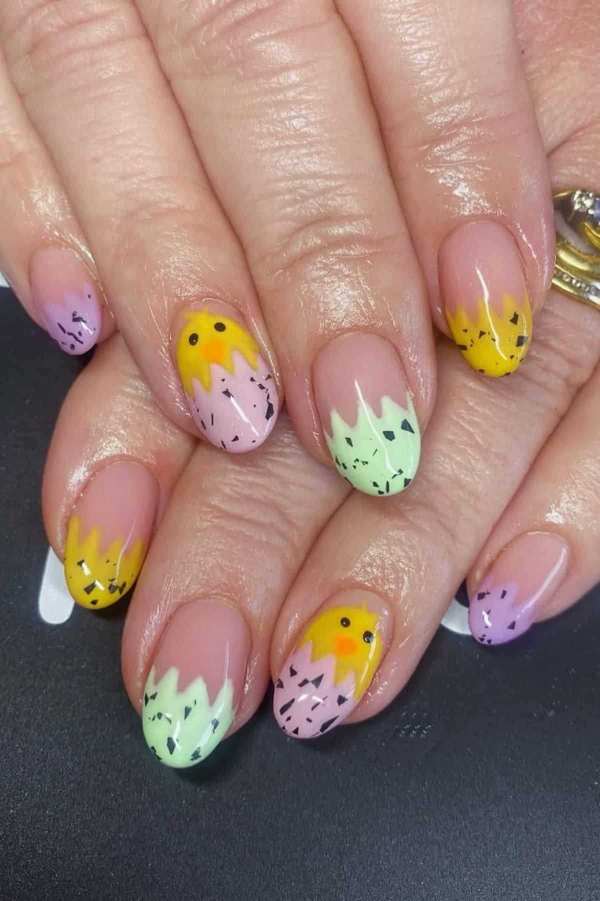 Close-up of hands with almond-shaped nails featuring pastel purple, yellow, and green jagged French tips with black speckles and small black smiley faces on some nails.
